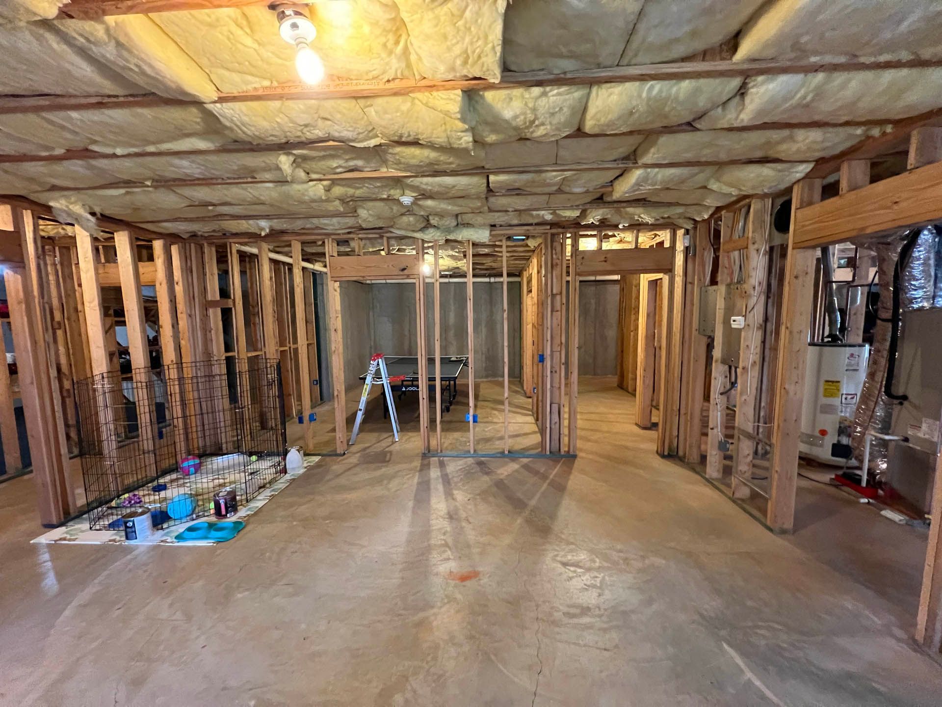 Basement under renovation with exposed wooden framing, insulation, and a concrete floor.  A ladder stands in the distance.