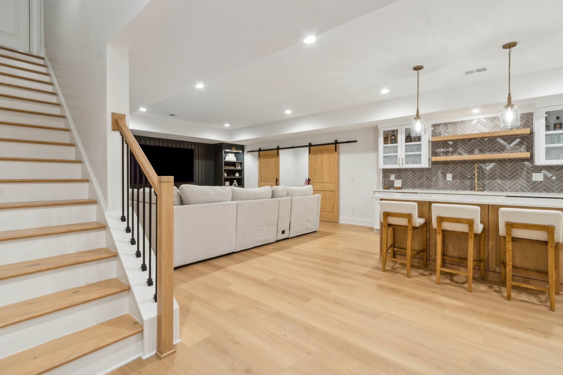 A bright, finished basement with a kitchen, living area, and staircase featuring light wood floors and neutral tones.
