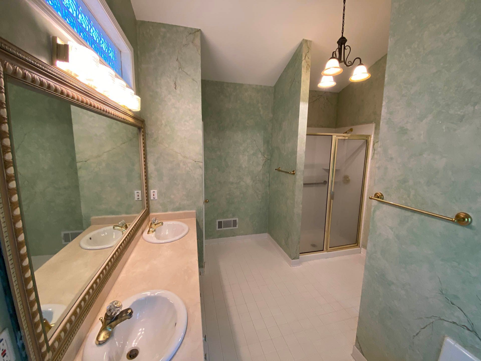A bathroom with green textured walls, a large mirror, dual sinks, and a shower with a frosted glass door. The floor is white.