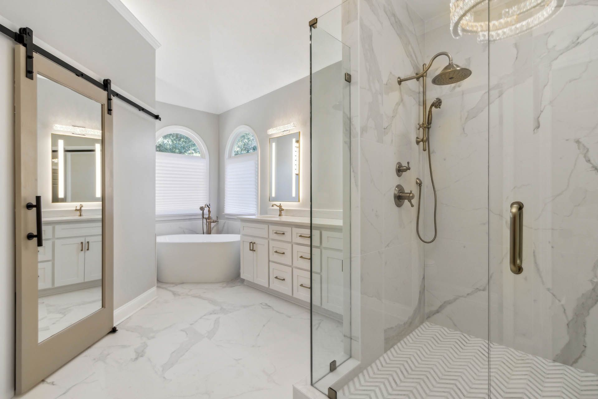 Spacious, bright bathroom with white marble flooring and walls. Features include a glass shower, soaking tub, and light fixtures with gold accents.