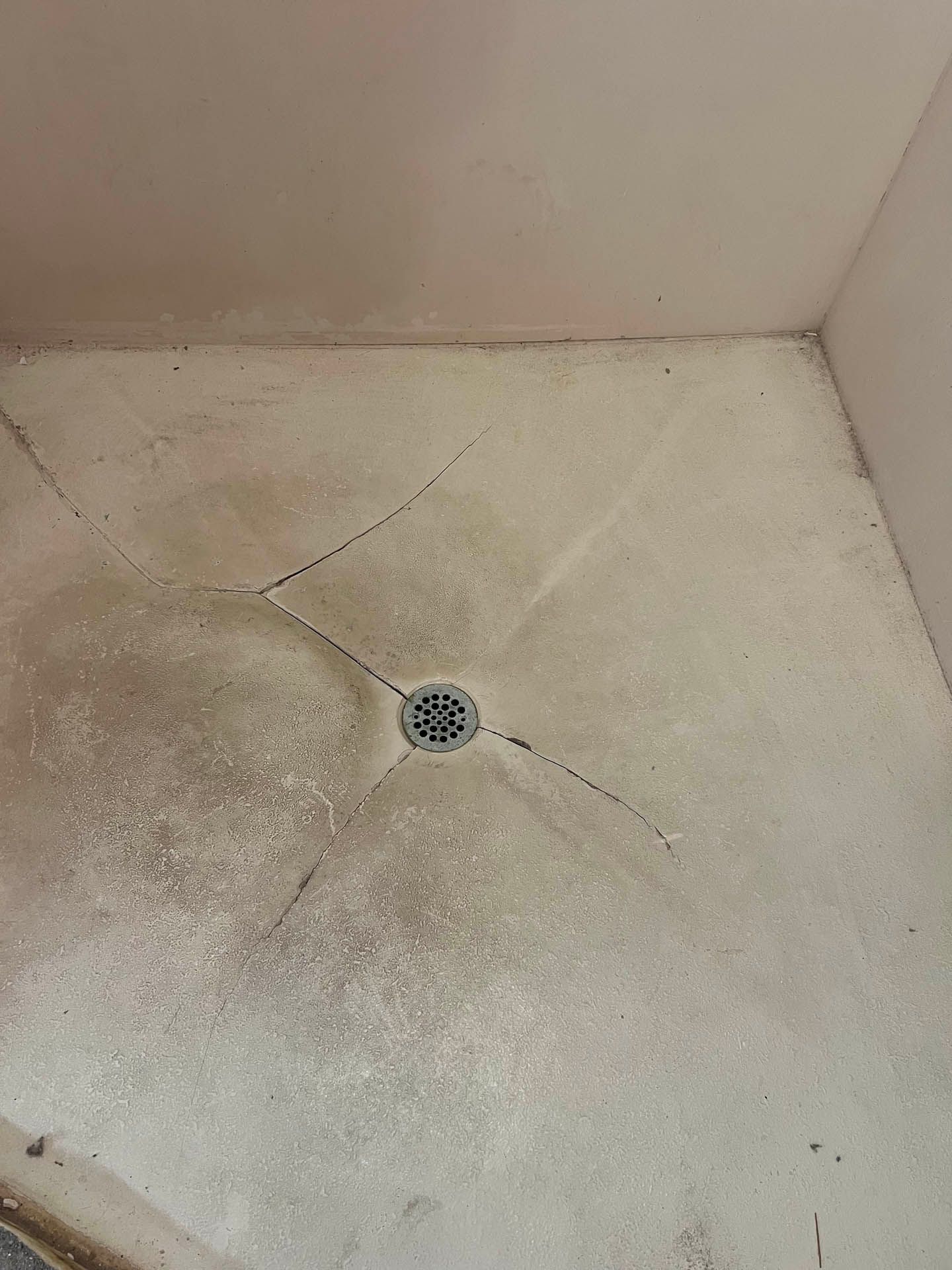 A shower stall floor with spiderweb-like cracks and a drain, appearing stained with discoloration and dirt.