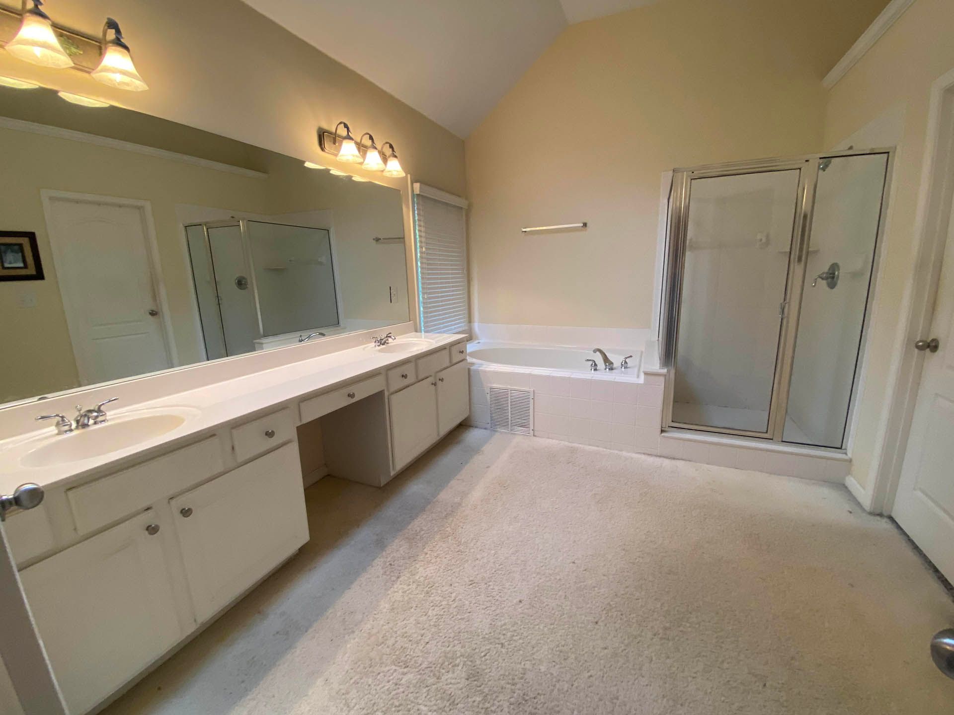 A spacious, well-lit bathroom with a large vanity, a glass shower, and a bathtub. The room has a beige color scheme and beige carpeting.