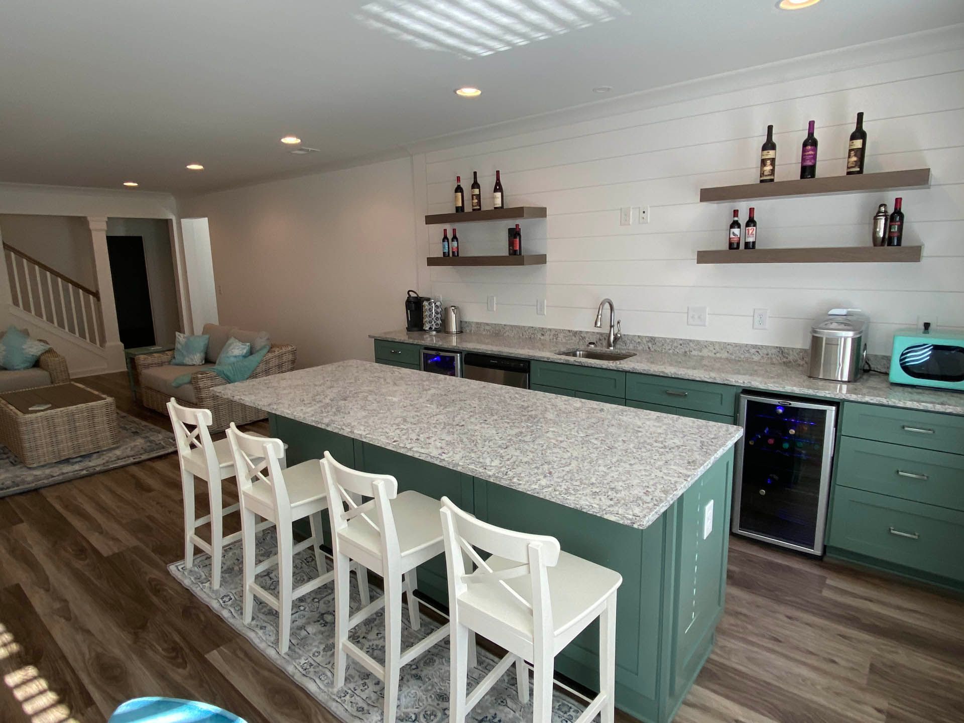 A basement bar area with a green island, white chairs, and shelves holding wine bottles. There's a sink, countertop, and additional seating visible.