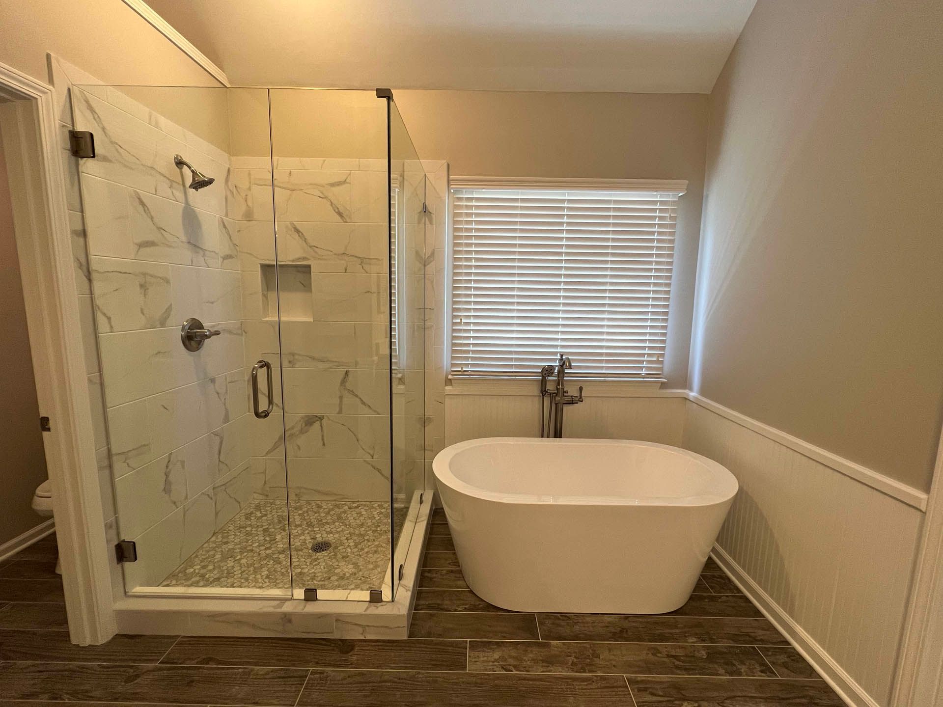A modern bathroom with a marble shower, freestanding bathtub, and window with blinds. Brown floor and tan walls.