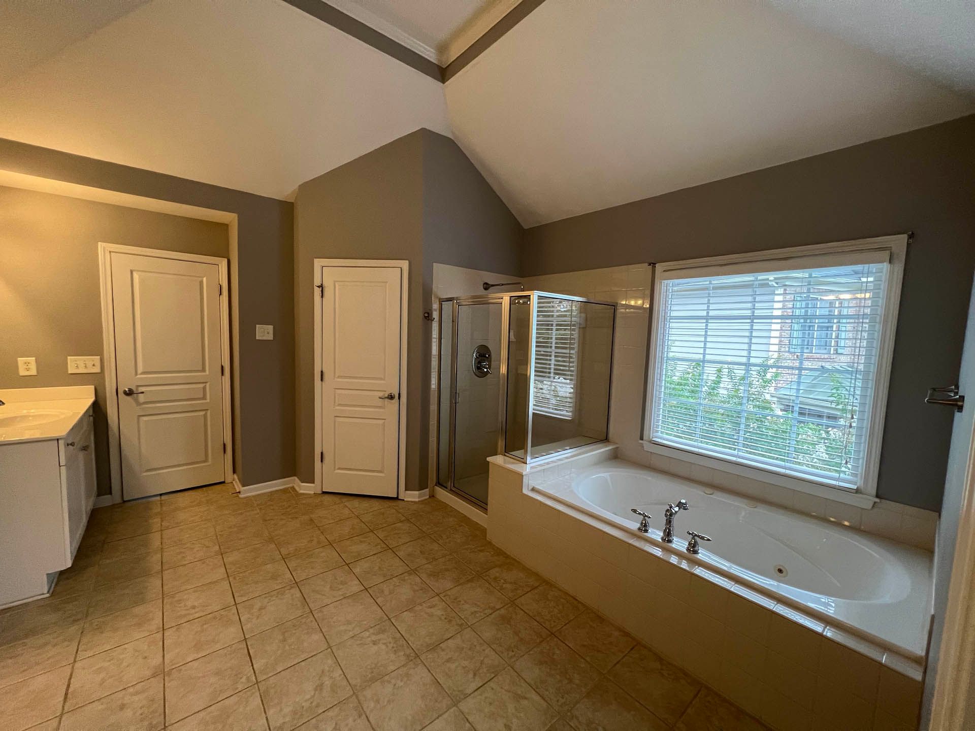 Spacious bathroom with a bathtub, shower, and two white doors. Beige tile floor and gray walls.