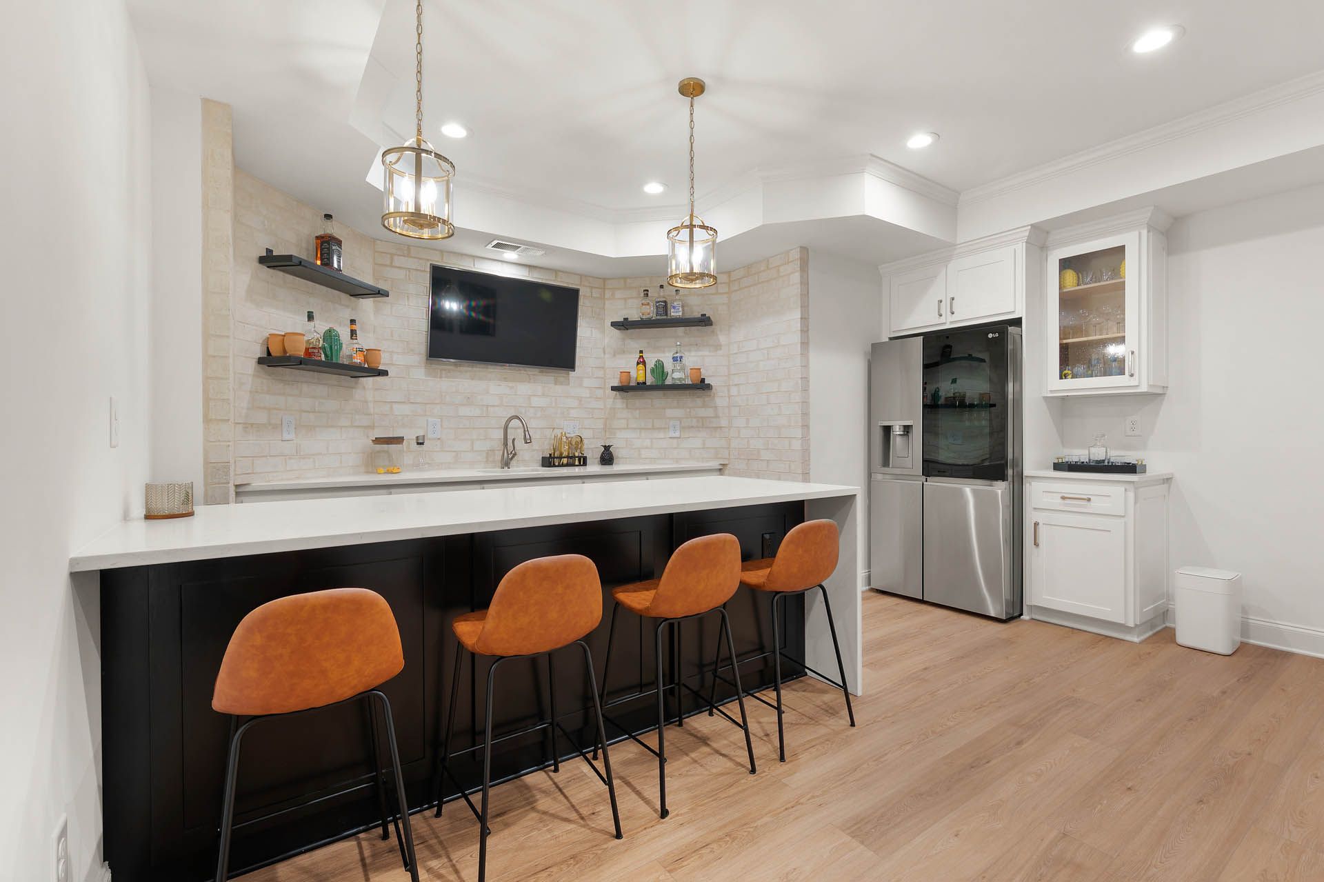 A modern basement bar with black and white surfaces, orange bar stools, and pendant lights. Includes a fridge, shelves, and a TV.