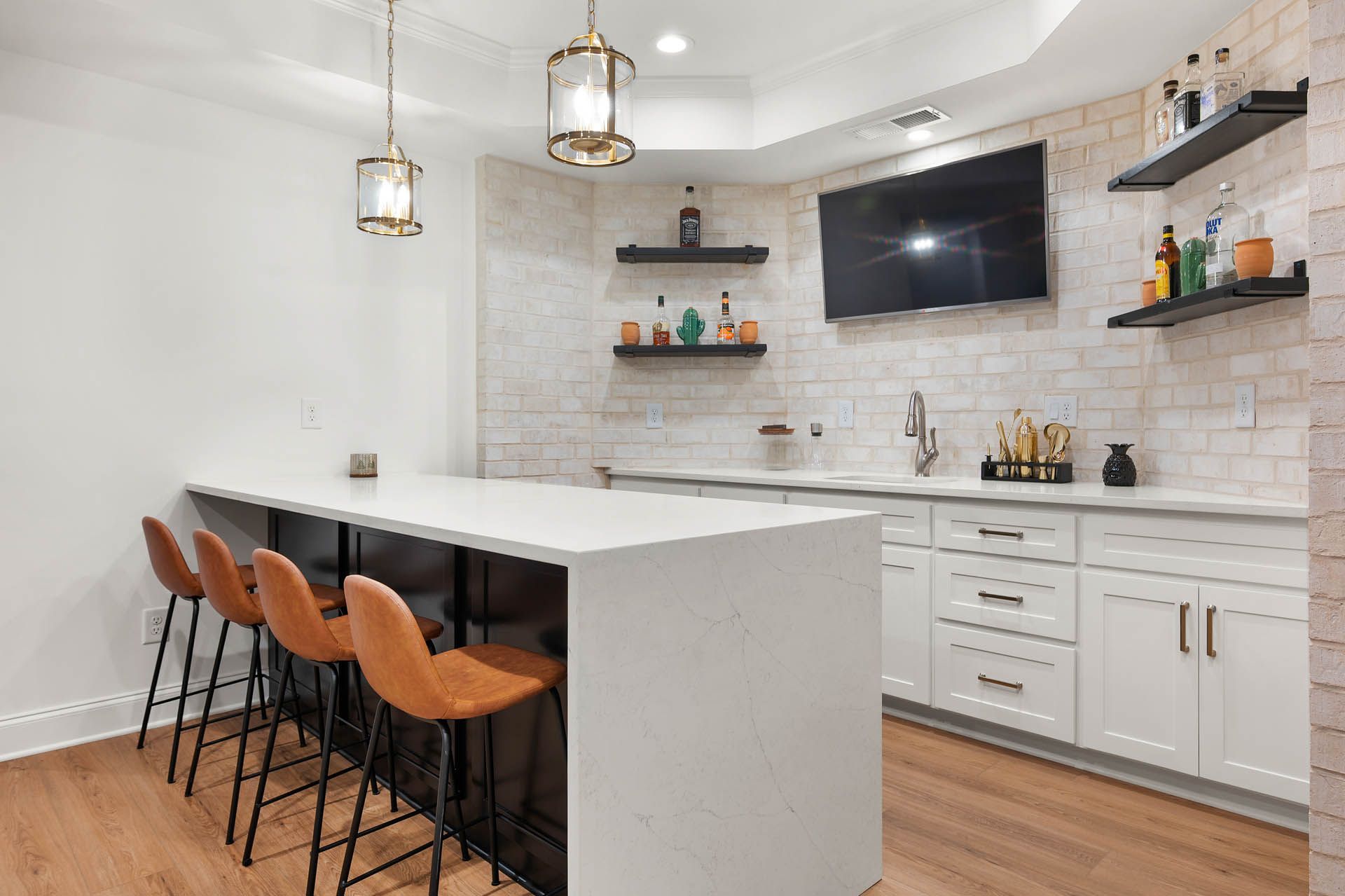 A modern home bar with a white countertop, brown bar stools, and a brick wall. Shelves display liquor bottles, with a TV and sink area.