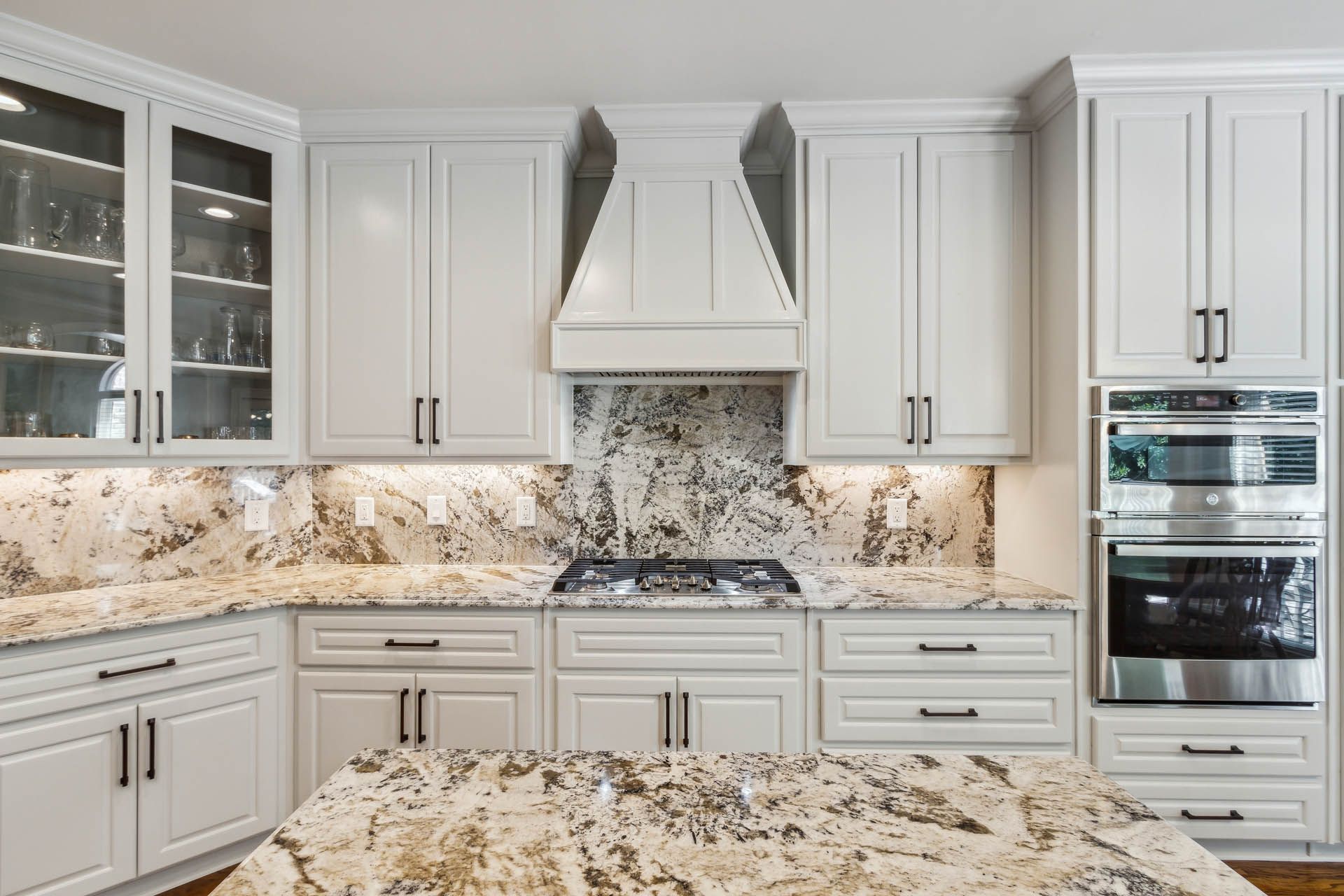 A bright kitchen with white cabinets, granite countertops, and stainless steel appliances. Includes a central island and range hood.