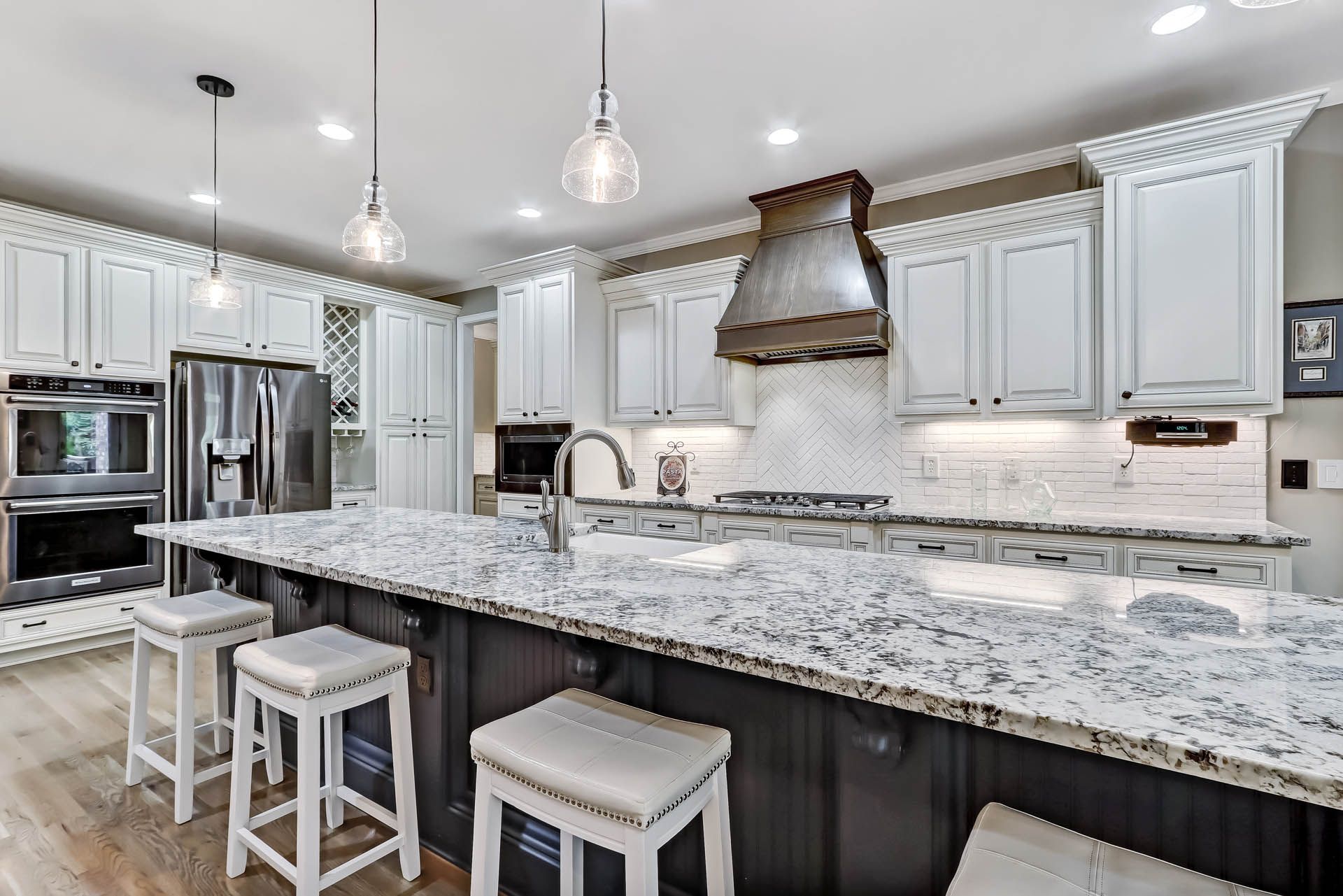 A bright, modern kitchen with white cabinets, a large granite island with stools, and stainless steel appliances.