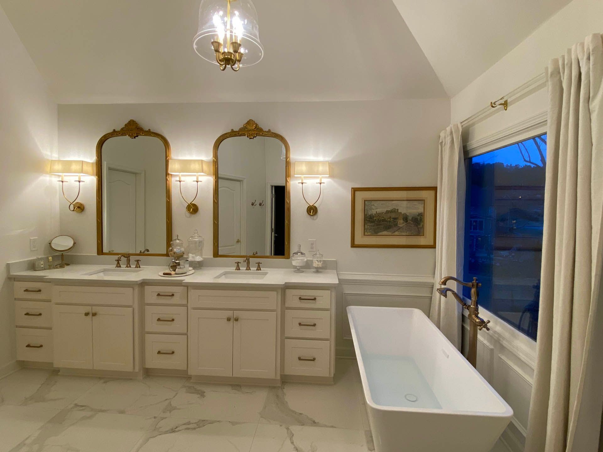 Luxurious white bathroom with a double vanity, ornate gold mirrors, a freestanding tub, and a window with draped curtains.