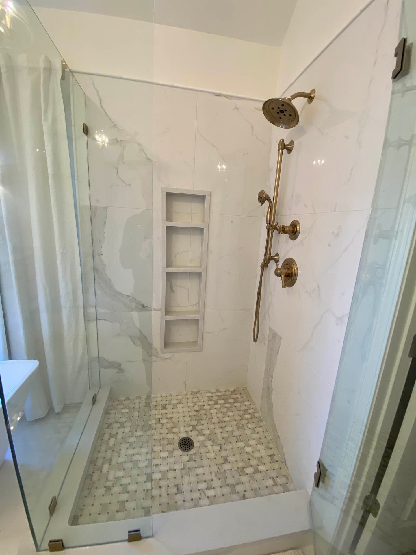 Modern bathroom shower with marble-look tile, pebble floor, glass doors, and gold fixtures. White curtain visible.