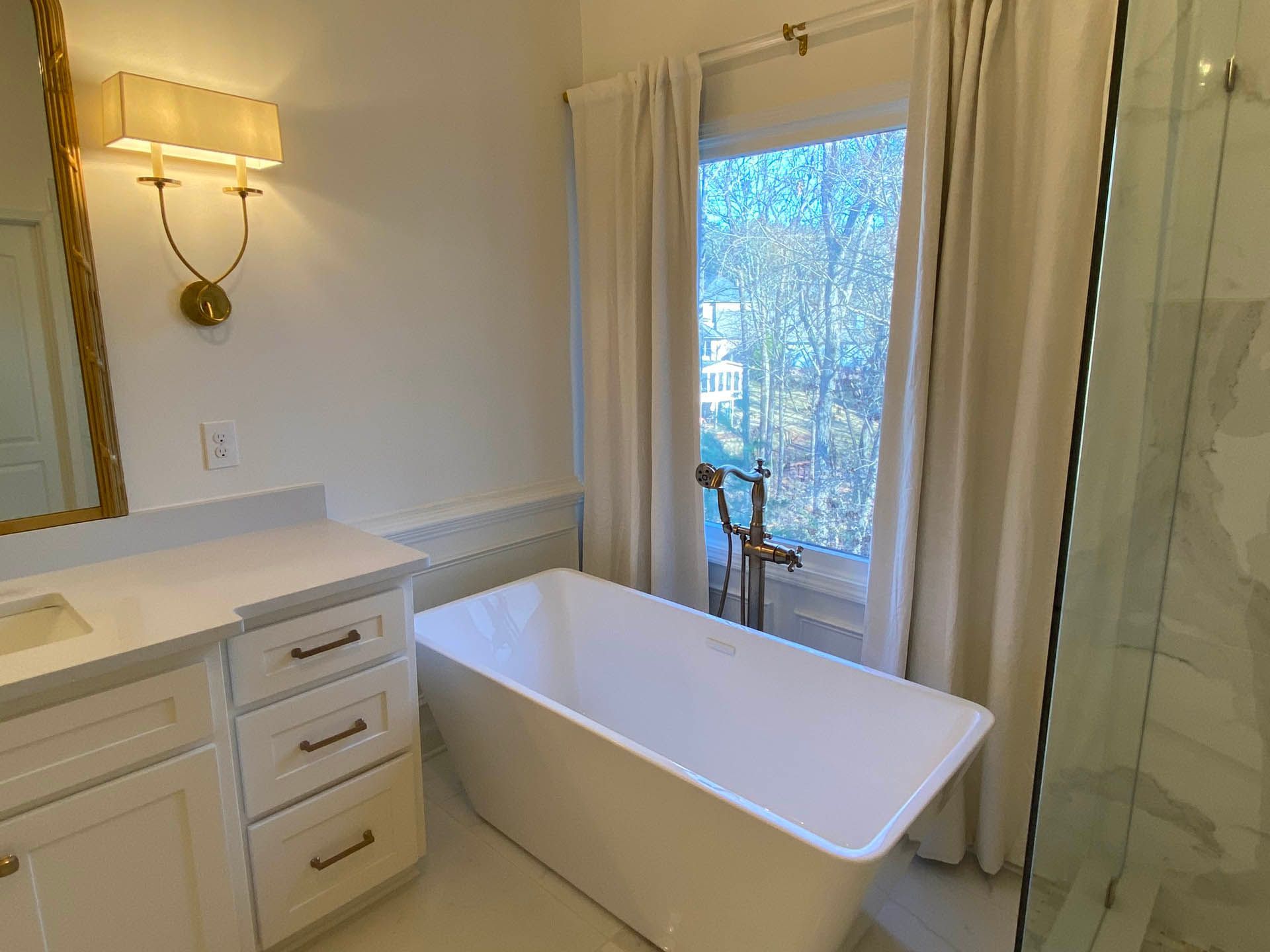A bright white bathroom with a freestanding bathtub, vanity, and window with white curtains. A gold sconce hangs on the wall.