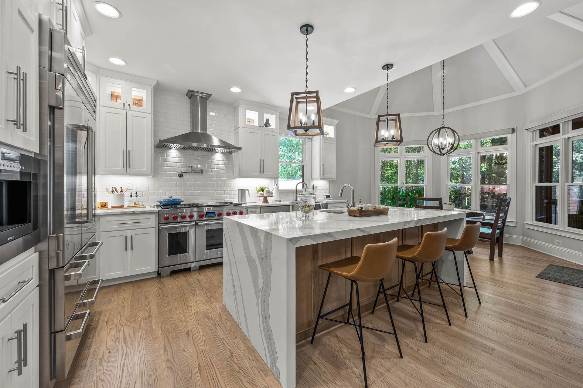 Spacious, bright kitchen with a large island, stainless steel appliances, white cabinets, and wooden floors. Bar stools line the island, and pendant lights hang overhead.