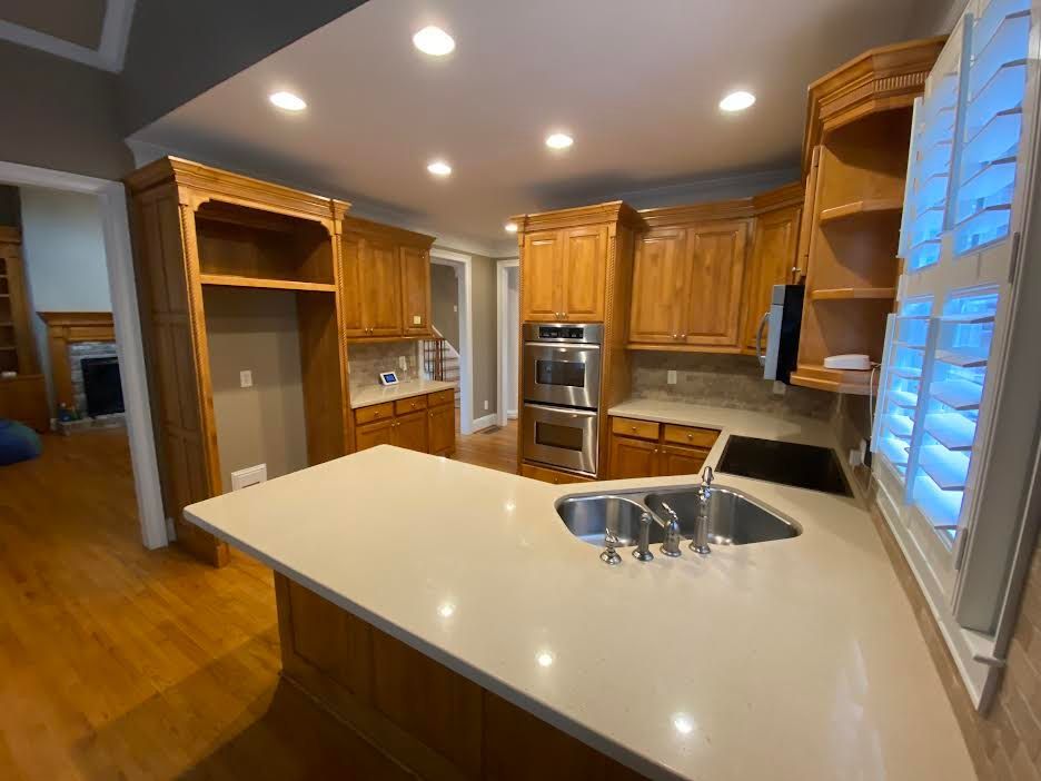 Spacious kitchen with light wooden cabinets, a light-colored countertop, and a stainless steel sink. Wooden floors and window shutters are also visible.