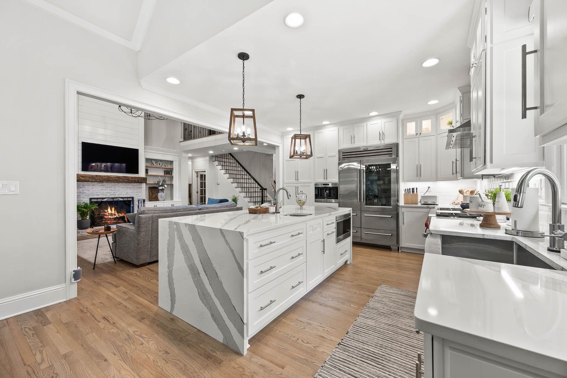 Spacious, modern kitchen with white cabinets, large island, stainless steel appliances, and hardwood floors. Open to a living area with a fireplace.