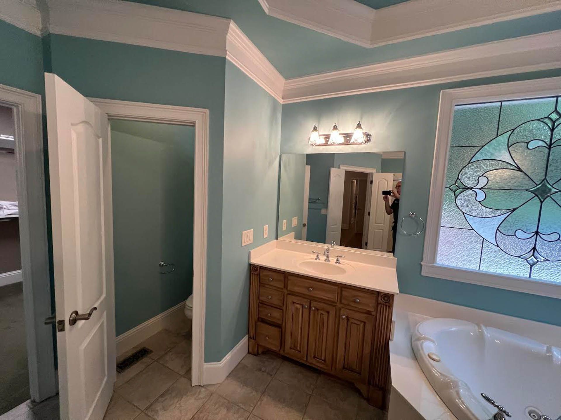 Bathroom with light blue walls, white trim, wooden vanity, and a stained glass window. A doorway leads to a separate toilet area.