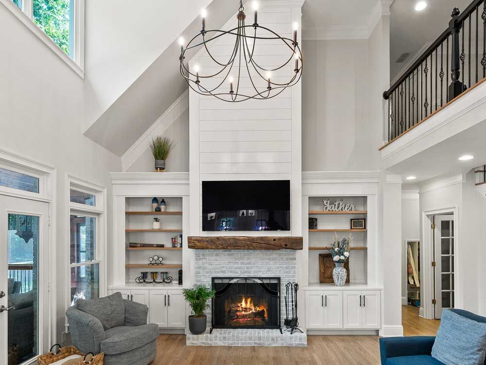 Living room with a fireplace, built-in shelves, a large chandelier, and a balcony. Bright white walls and wooden accents throughout.