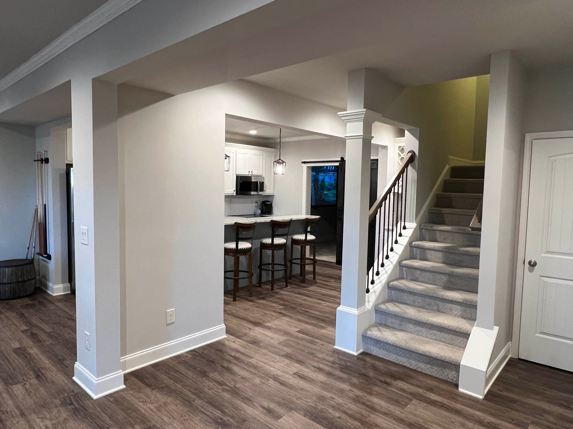 A renovated interior with a kitchen, stairs, and columns, featuring wood-look flooring, light gray walls, and white trim.