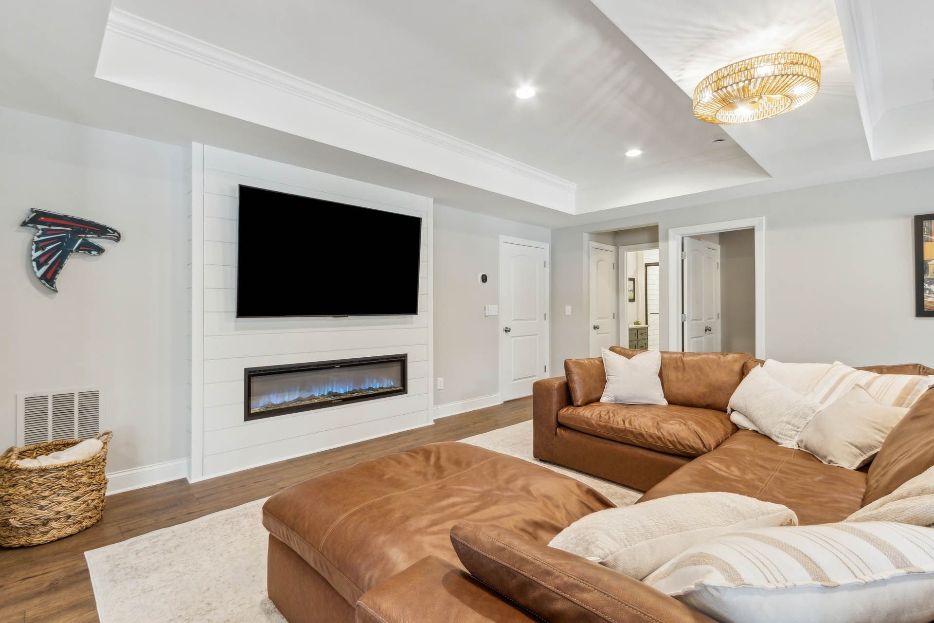 A modern living room with a brown leather sectional, fireplace, TV, and Atlanta Falcons logo. White trim, light-colored walls, and a rug create a bright space.