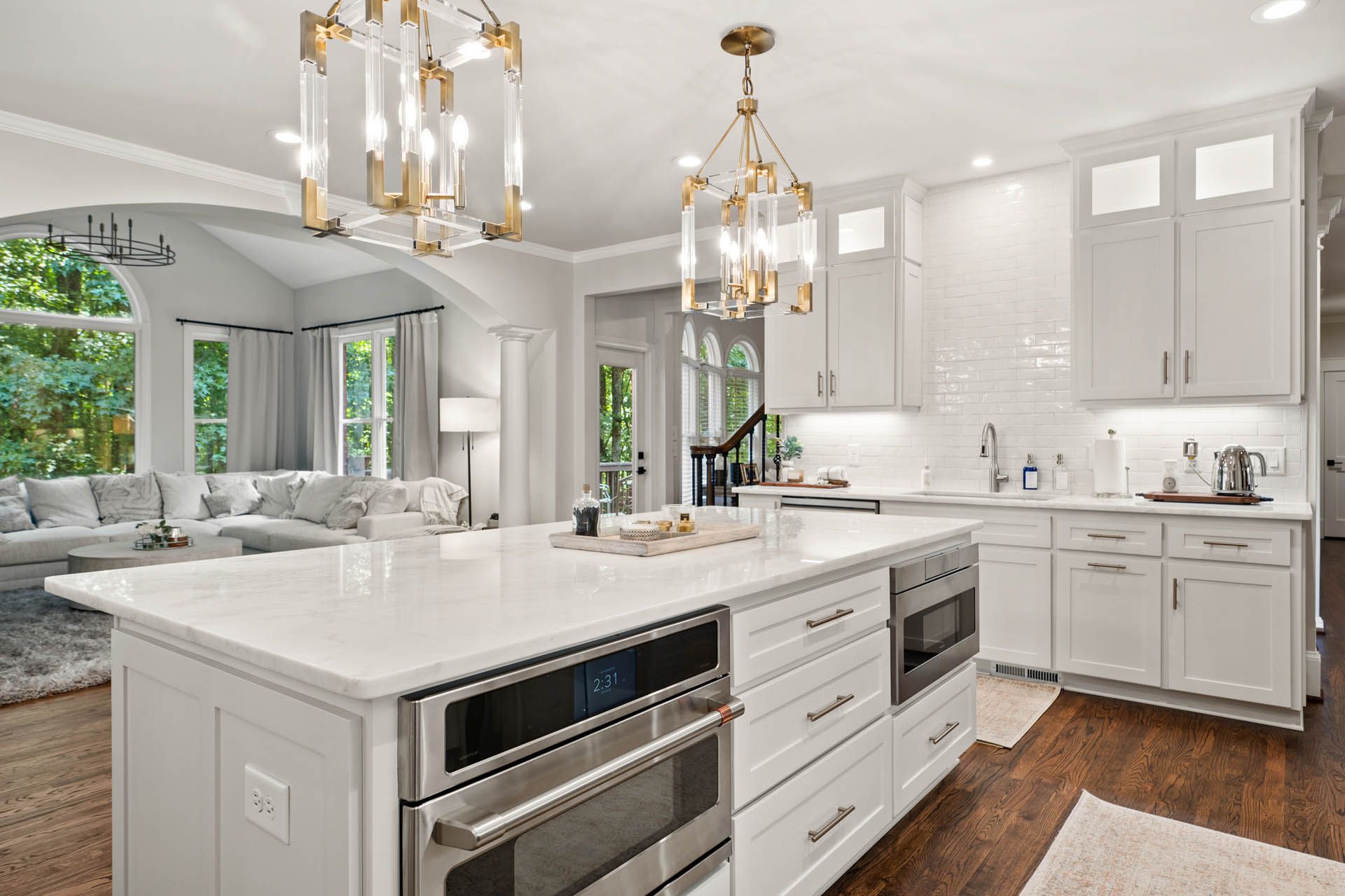 Bright white kitchen with an island, cabinets, and two gold chandeliers. A living room is visible in the background.