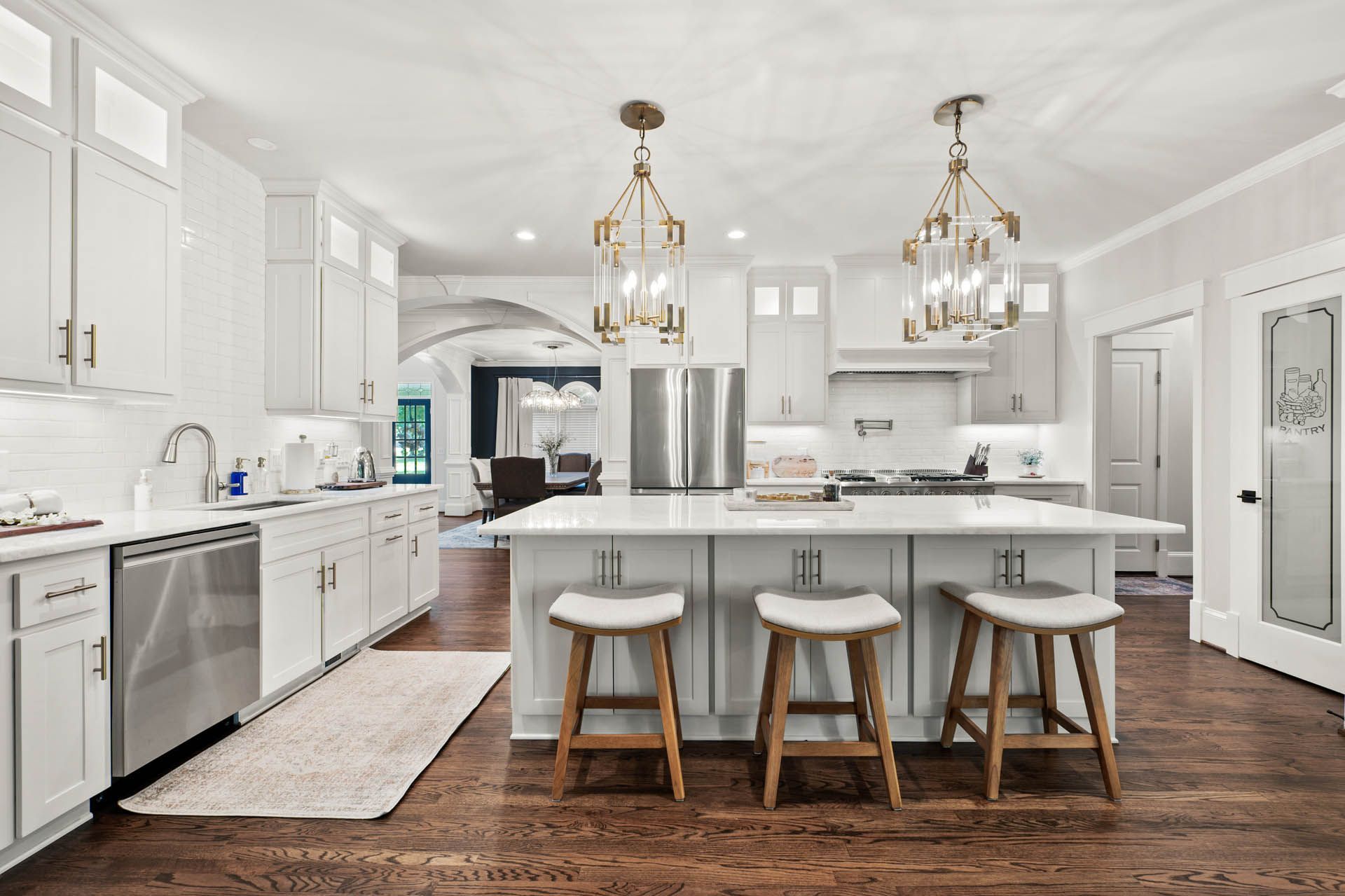 Bright white kitchen with stainless steel appliances, wooden stools at an island, and hardwood floors. Arched doorway leads to a dining area.