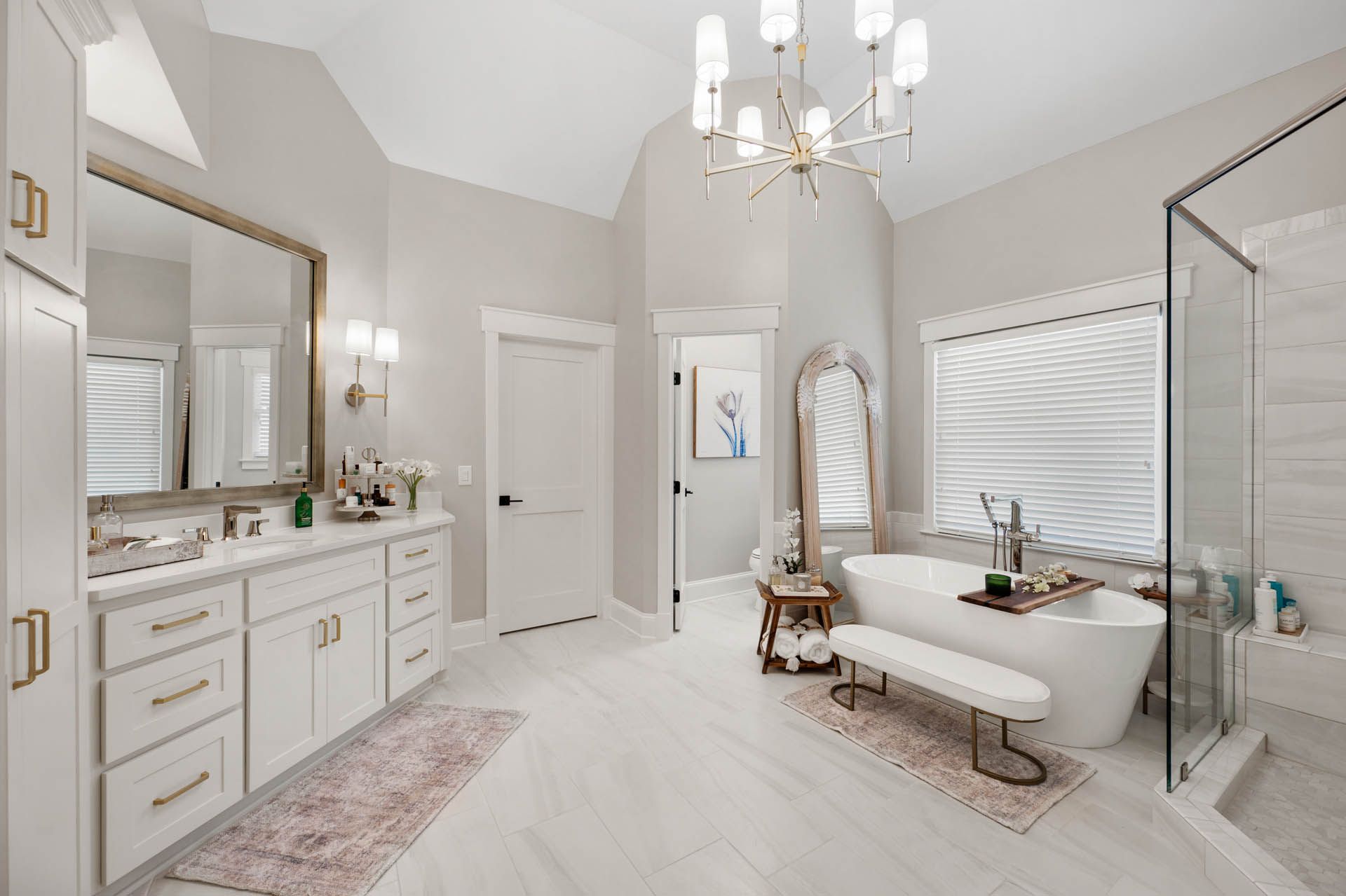 Spacious, bright bathroom with a white bathtub, shower, vanity, and chandelier. Neutral tones with touches of pink and gold.