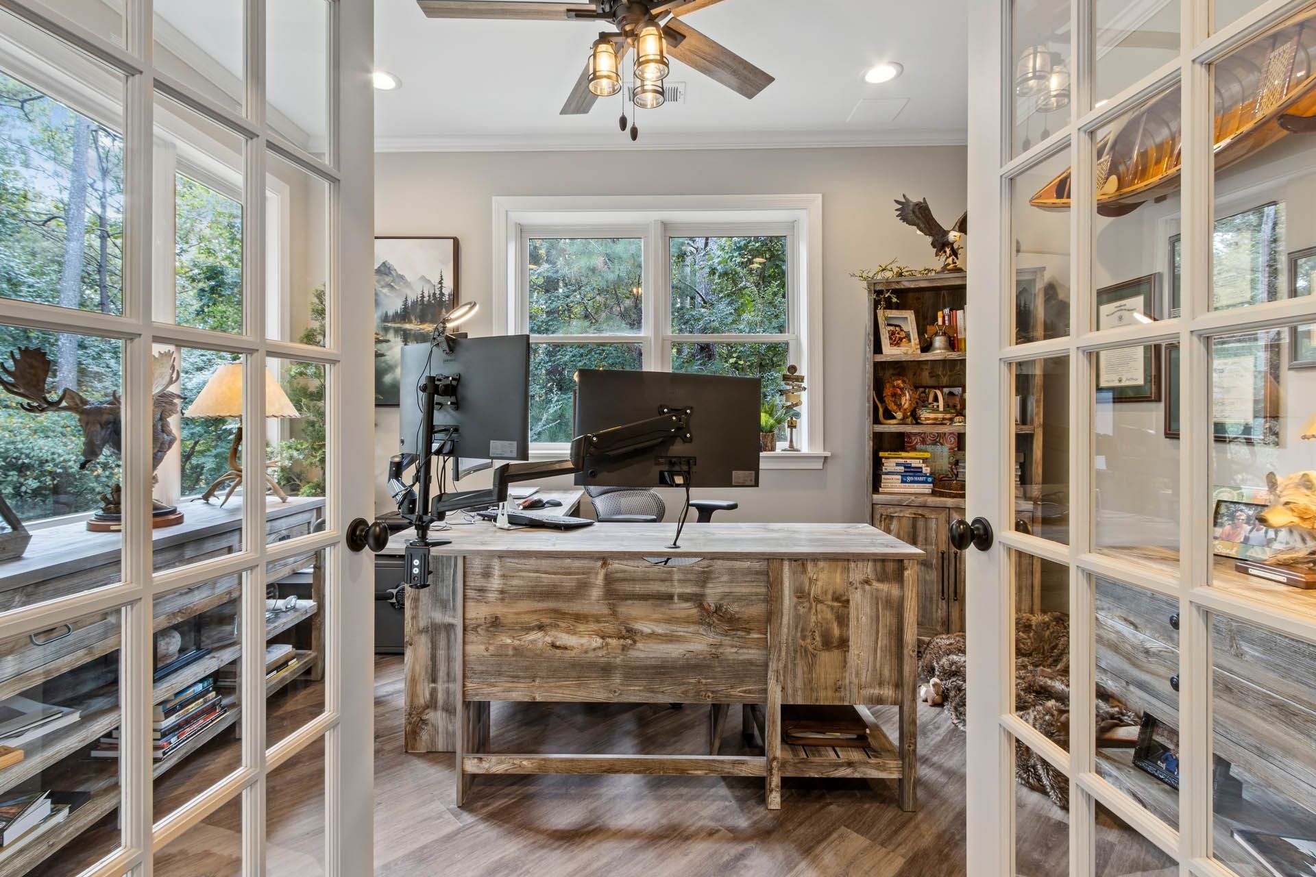 Home office with rustic wooden desk, two computer monitors, and bookshelves visible through French doors. Sunlight streams in from the windows.