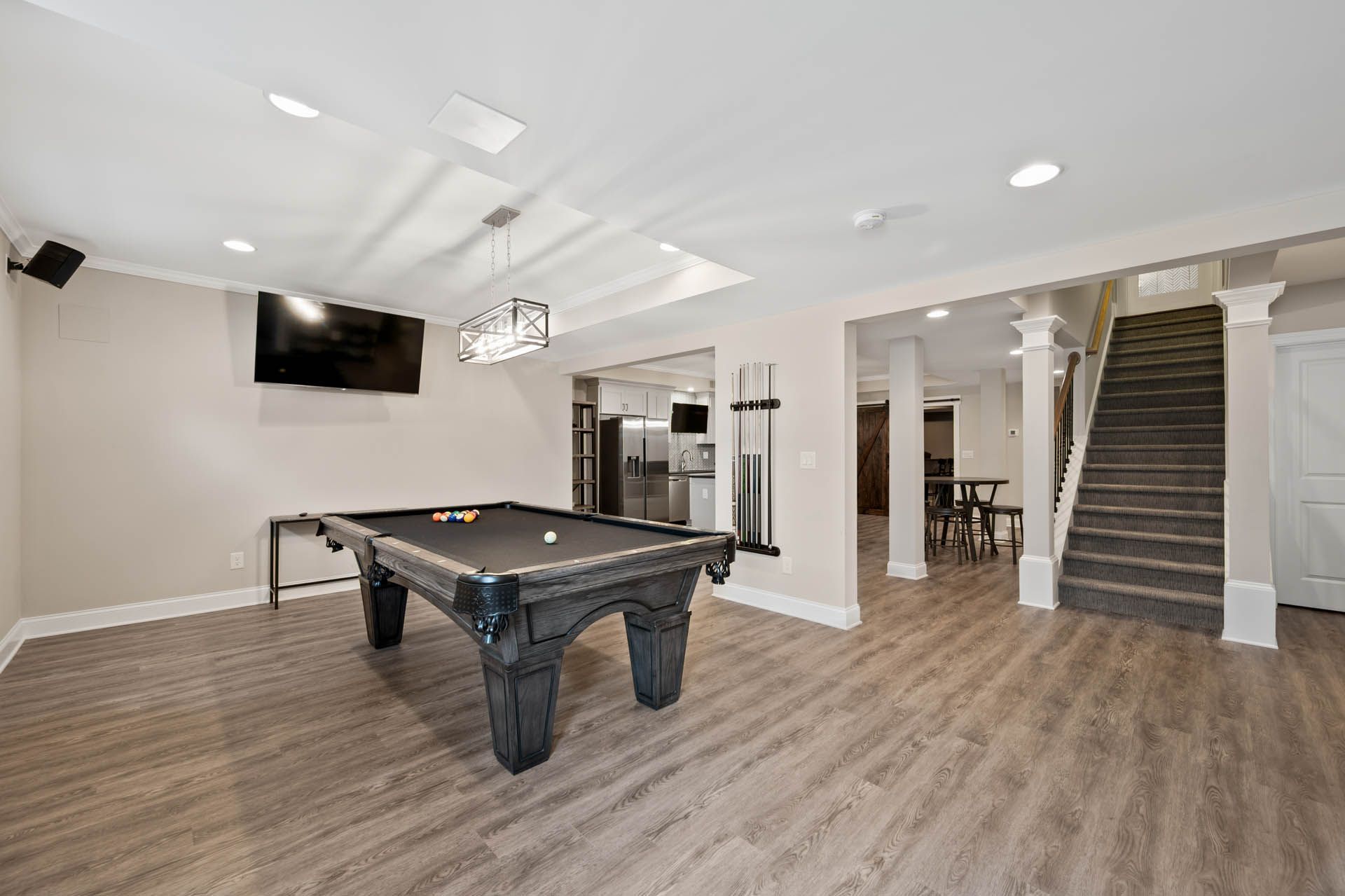 A finished basement with a pool table, TV, and stairs leading to another room with a kitchen and dining area.