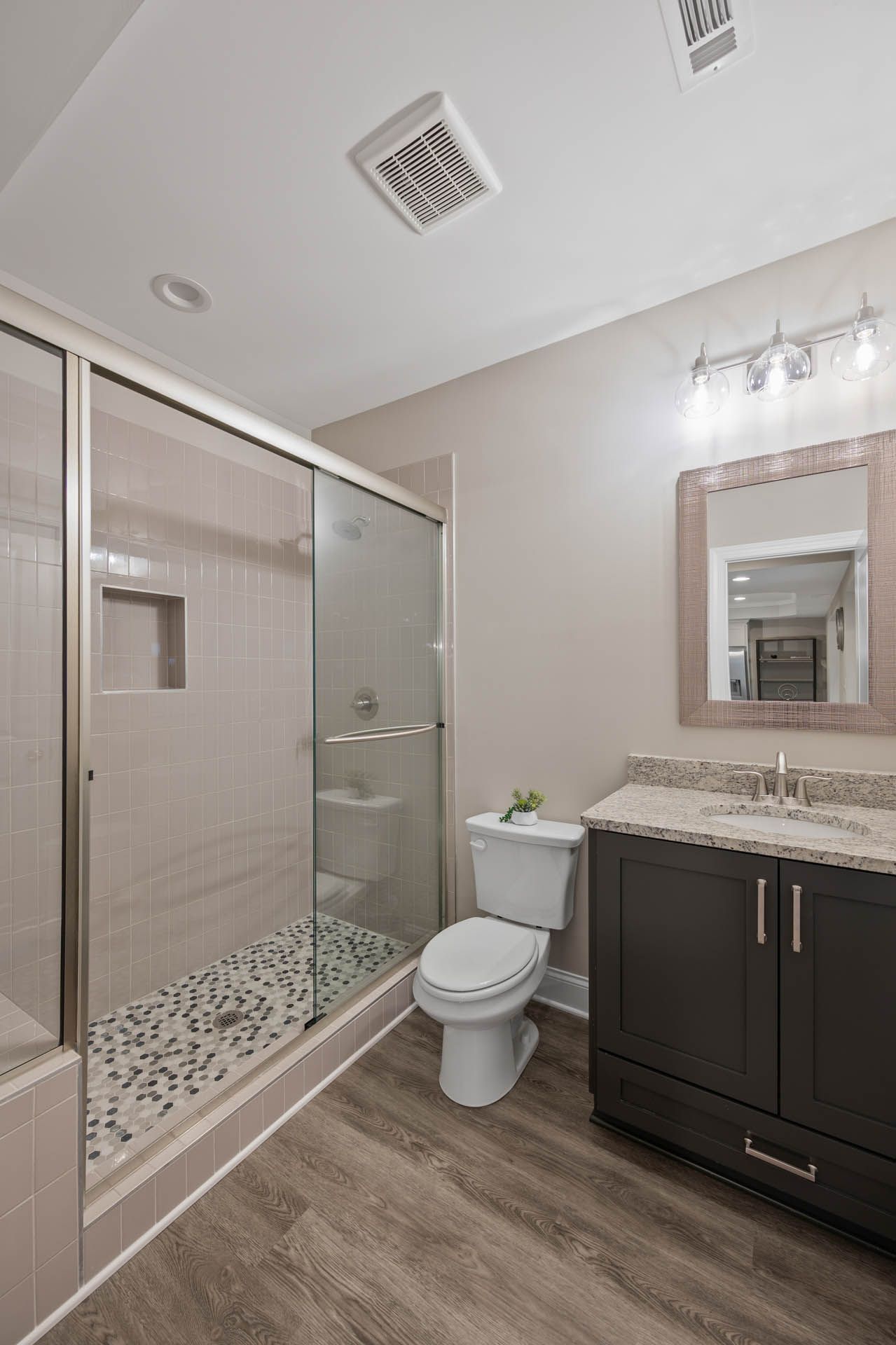 A modern bathroom with a glass shower, toilet, dark wood vanity, and stone-tiled shower floor. The walls are beige, and the floor is wood-look.