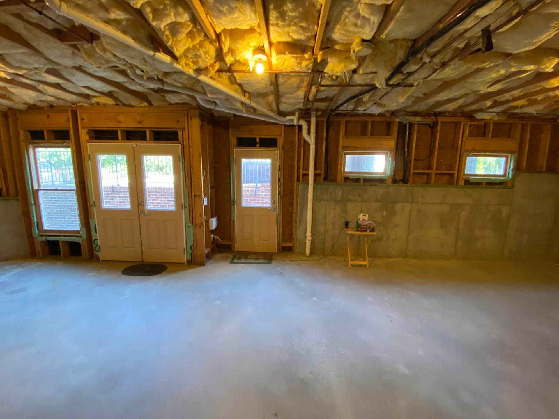 Unfinished basement with concrete floor, exposed beams, insulation, doorways, and small windows; a small table sits near the right wall.