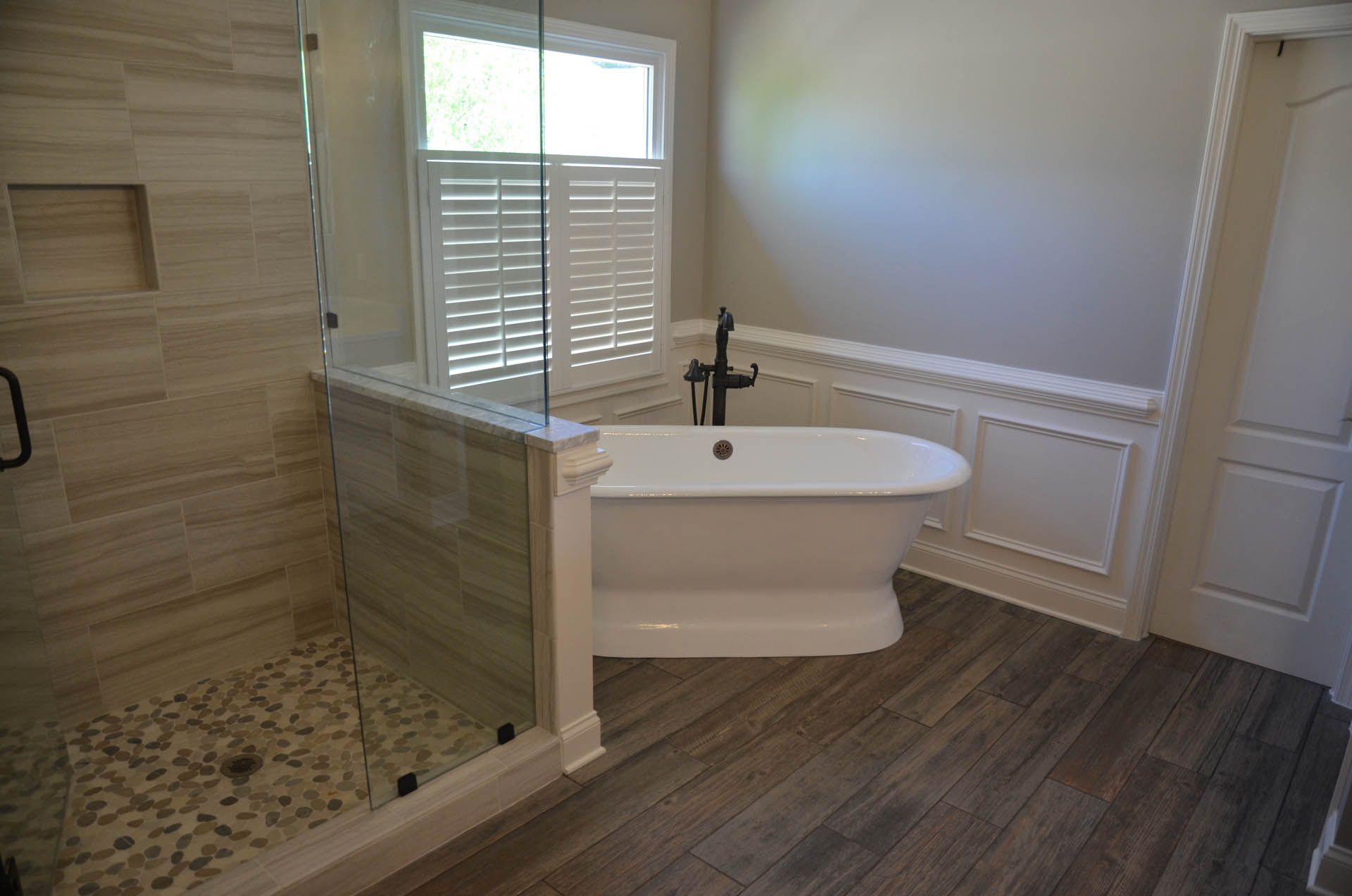 Spacious bathroom with a freestanding white bathtub, a glass-walled shower, and a window with white shutters. Wood-look tile flooring.