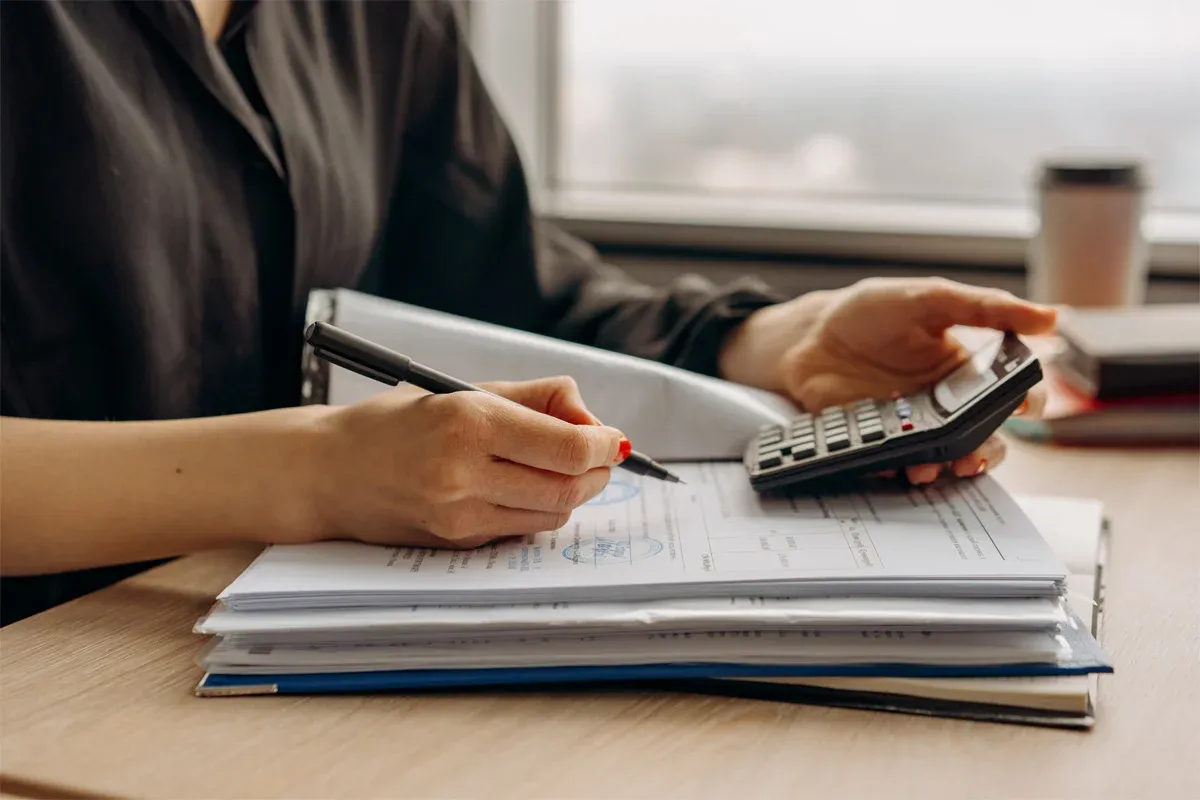 Person using a calculator, writing on papers, near a window with a coffee cup.