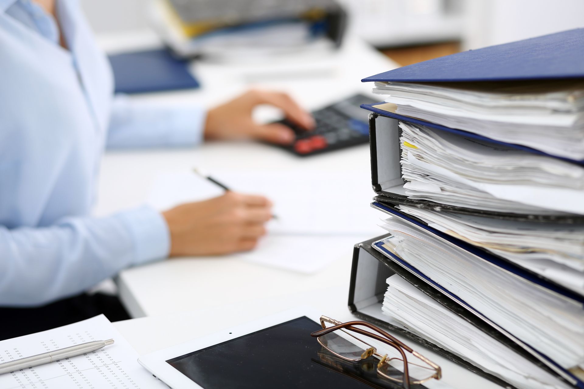 Person at desk with calculator and pen; a stack of binders and glasses in foreground.