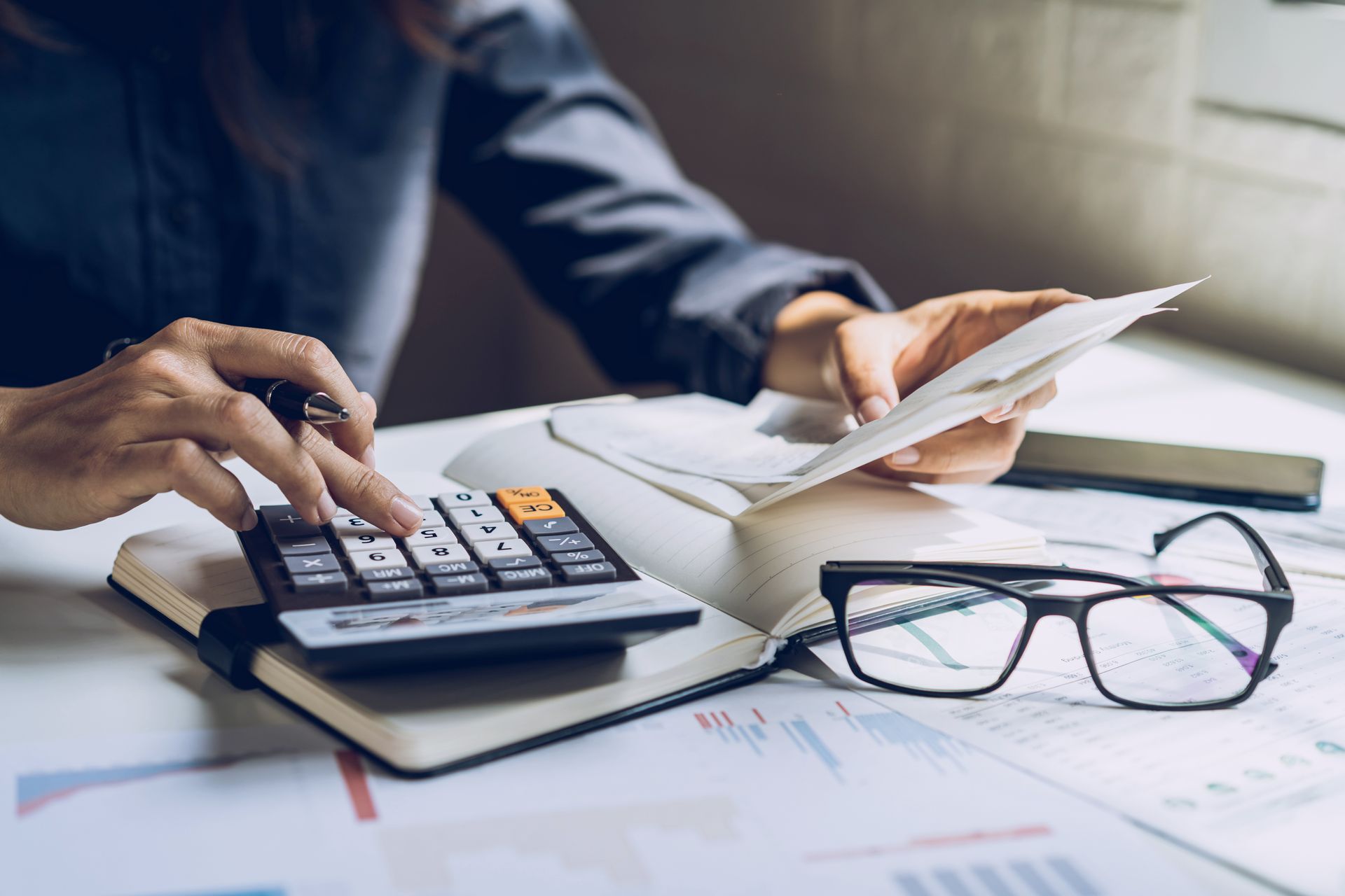 Person calculating expenses with a calculator, receipts, and glasses on a desk.