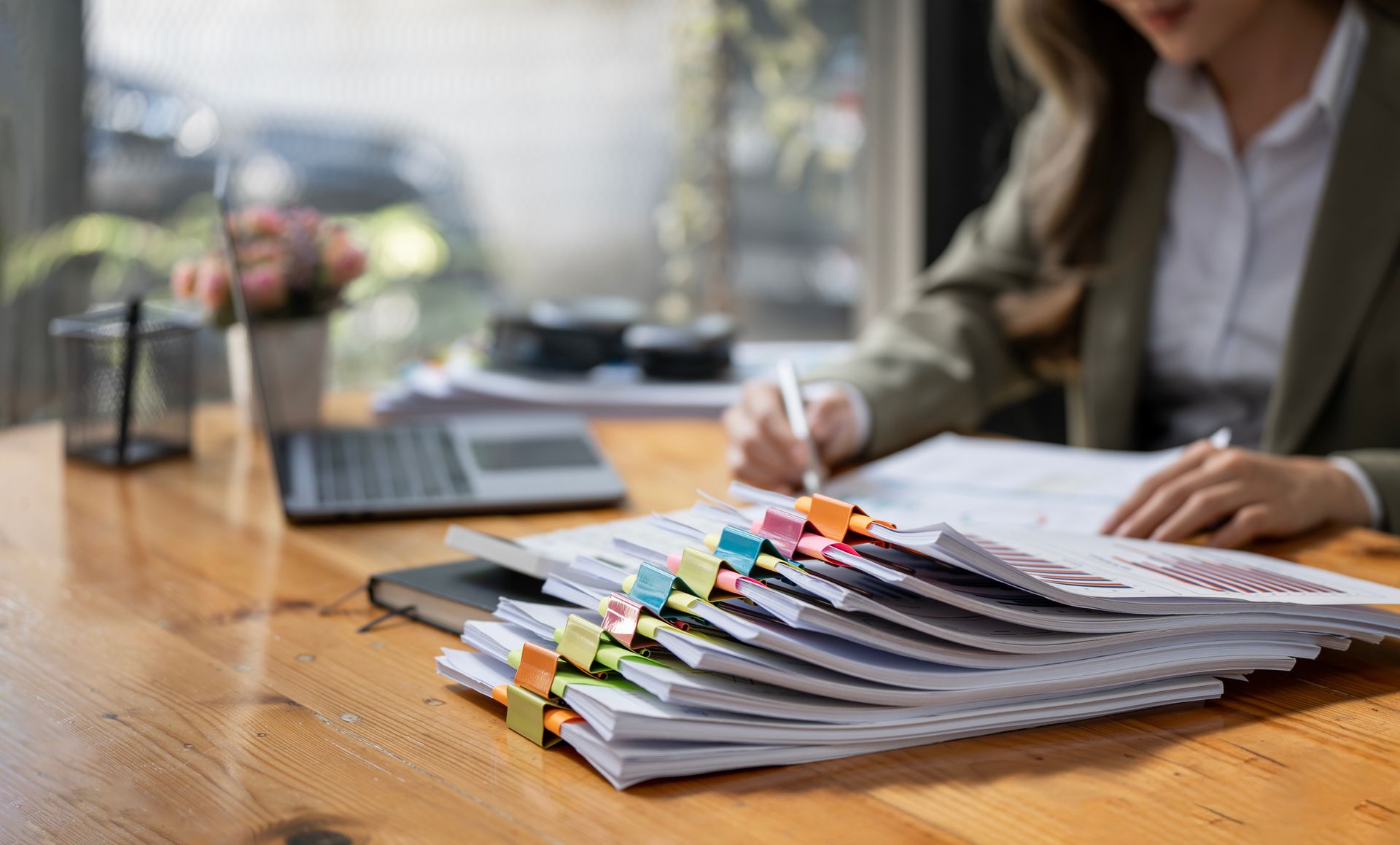 Woman in blazer writing at desk with stacked documents and laptop.