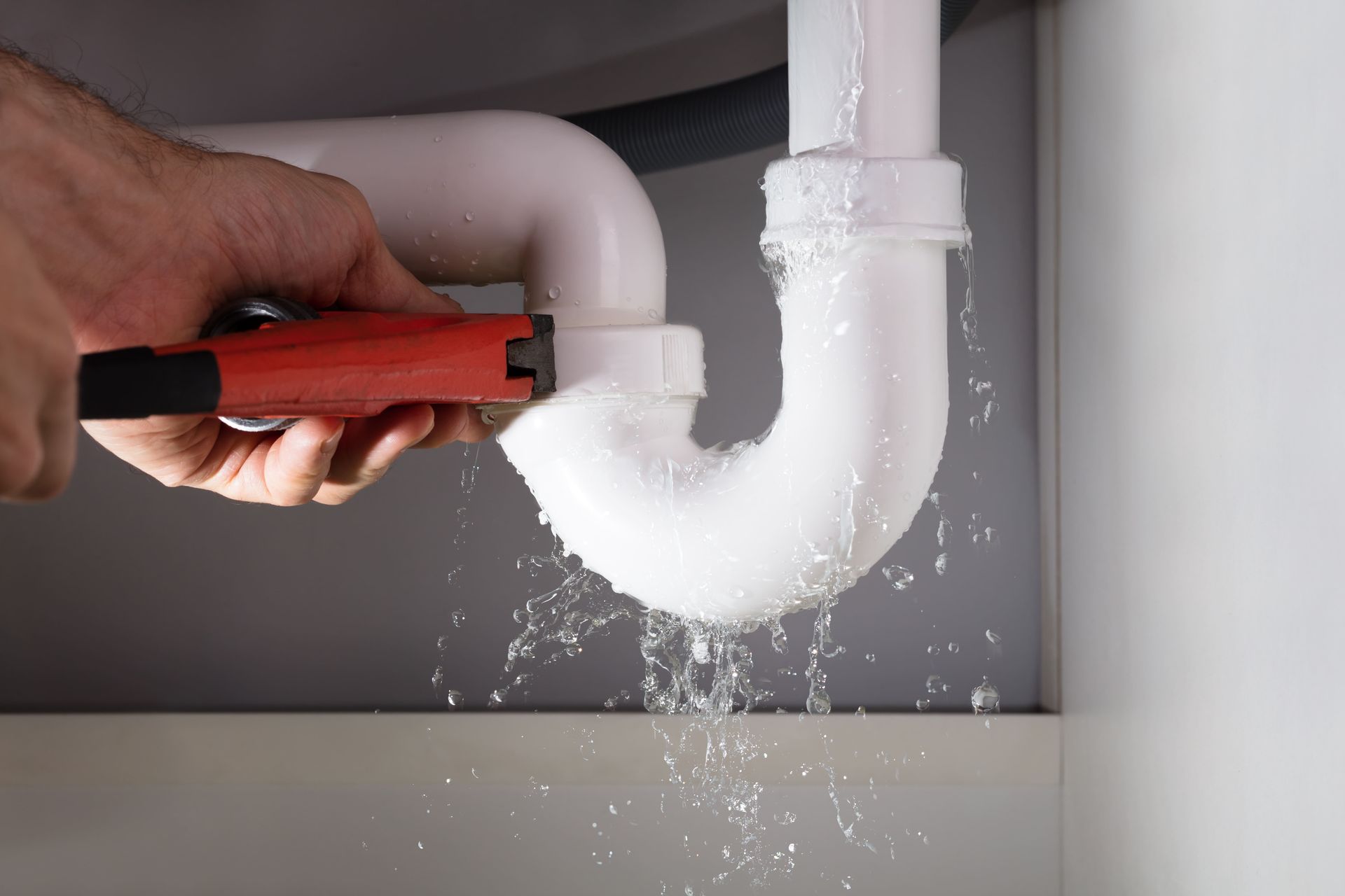 Hand tightening a pipe joint, water spraying in a cabinet; white pipes, red wrench, water droplets.