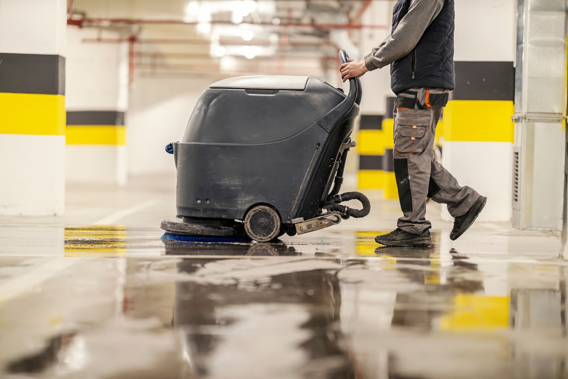 Worker pushing a floor scrubber in a parking garage with wet, reflective concrete floor