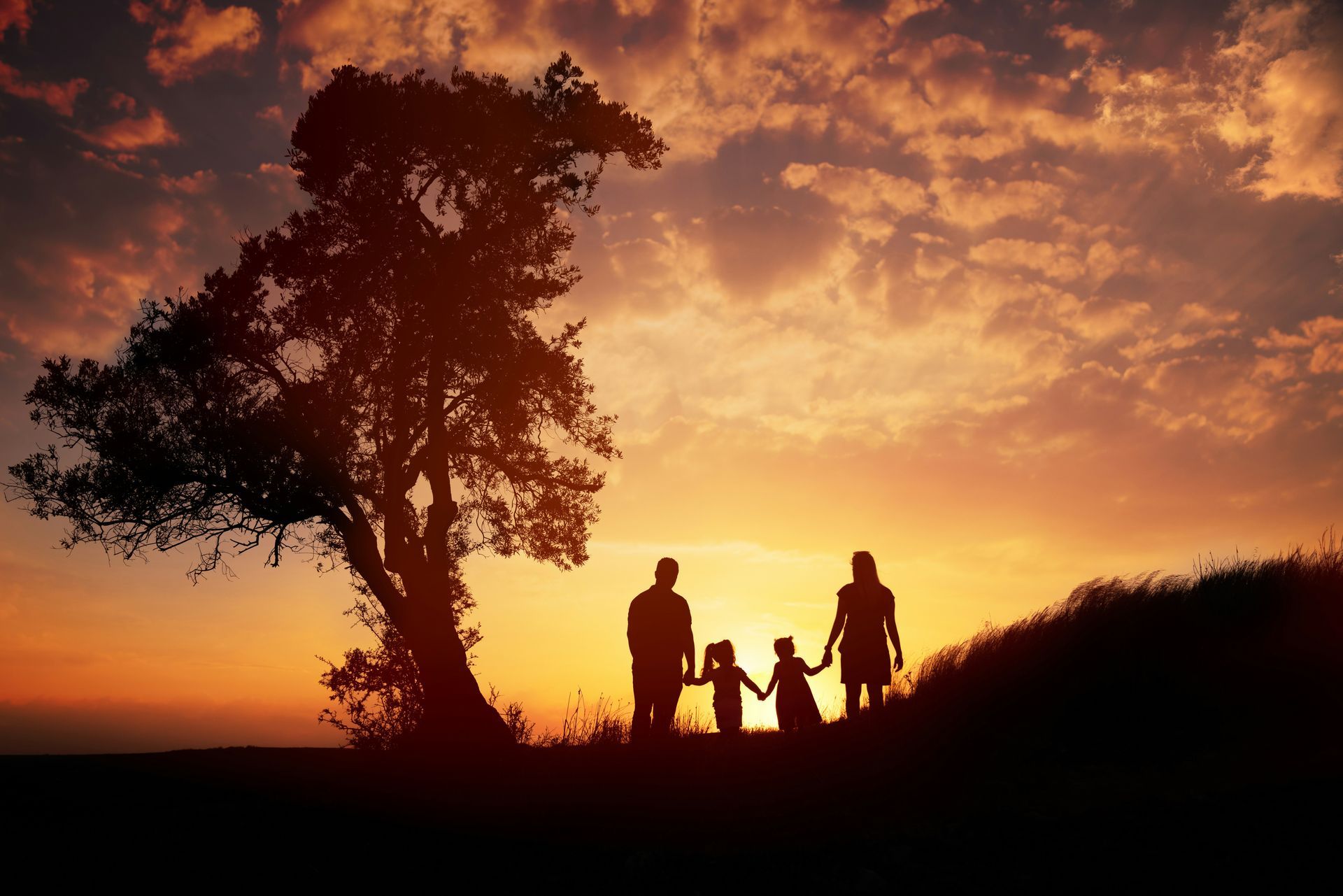 Family silhouette holding hands at sunset, with a tree on a hilltop.