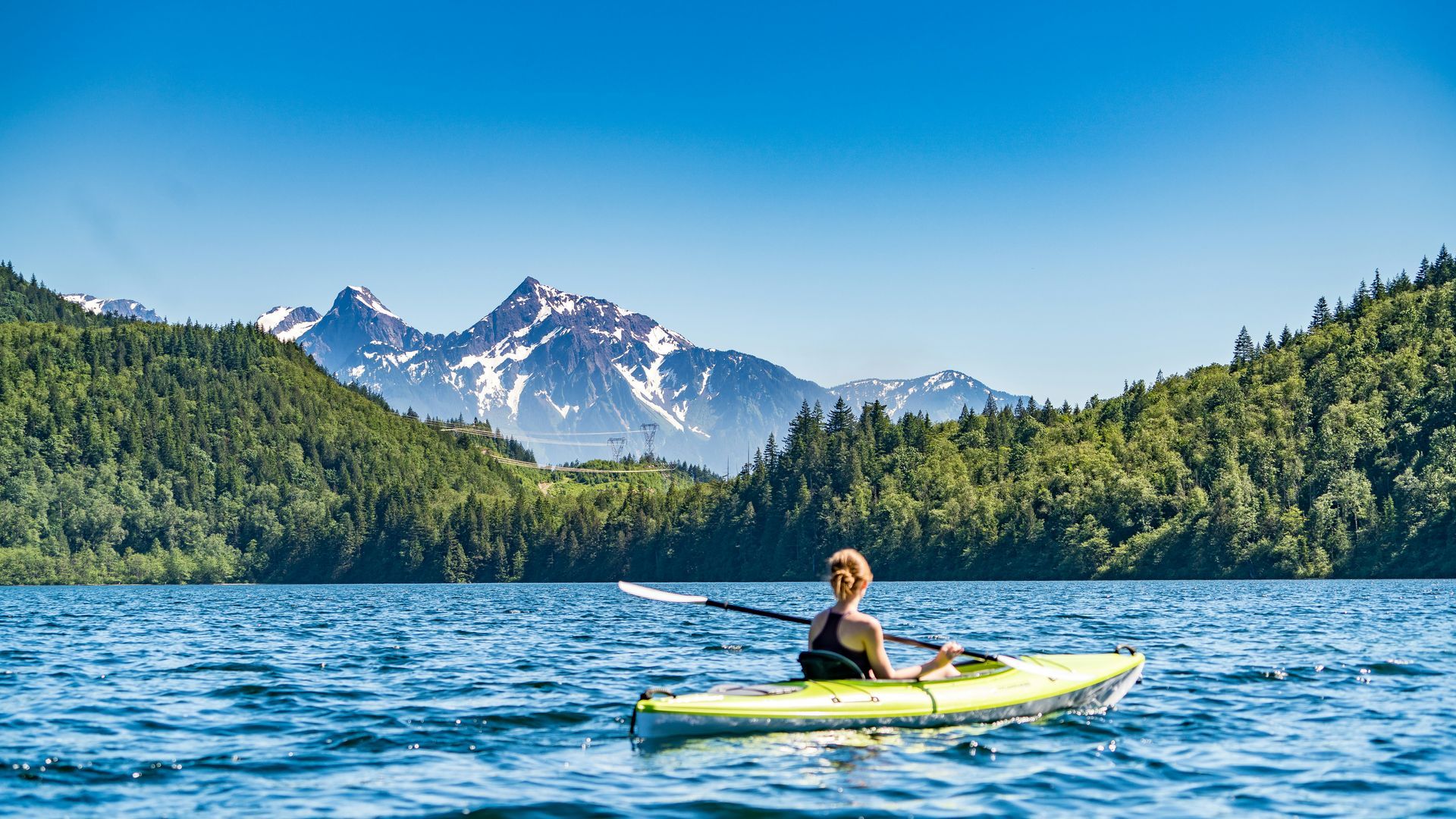 Person kayaking on a lake with mountains and trees in the background under a blue sky.