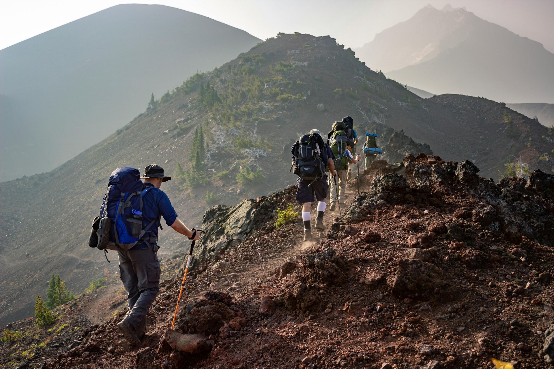 Hikers with backpacks ascend a rocky mountain ridge toward a distant peak in sunlight.