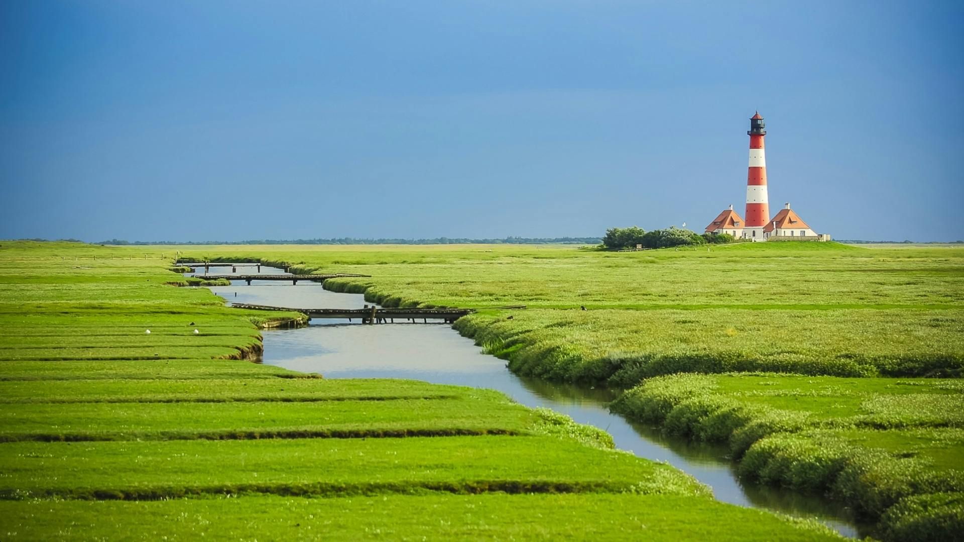 Lighthouse with red and white stripes beside a green marsh and waterway under a blue sky.