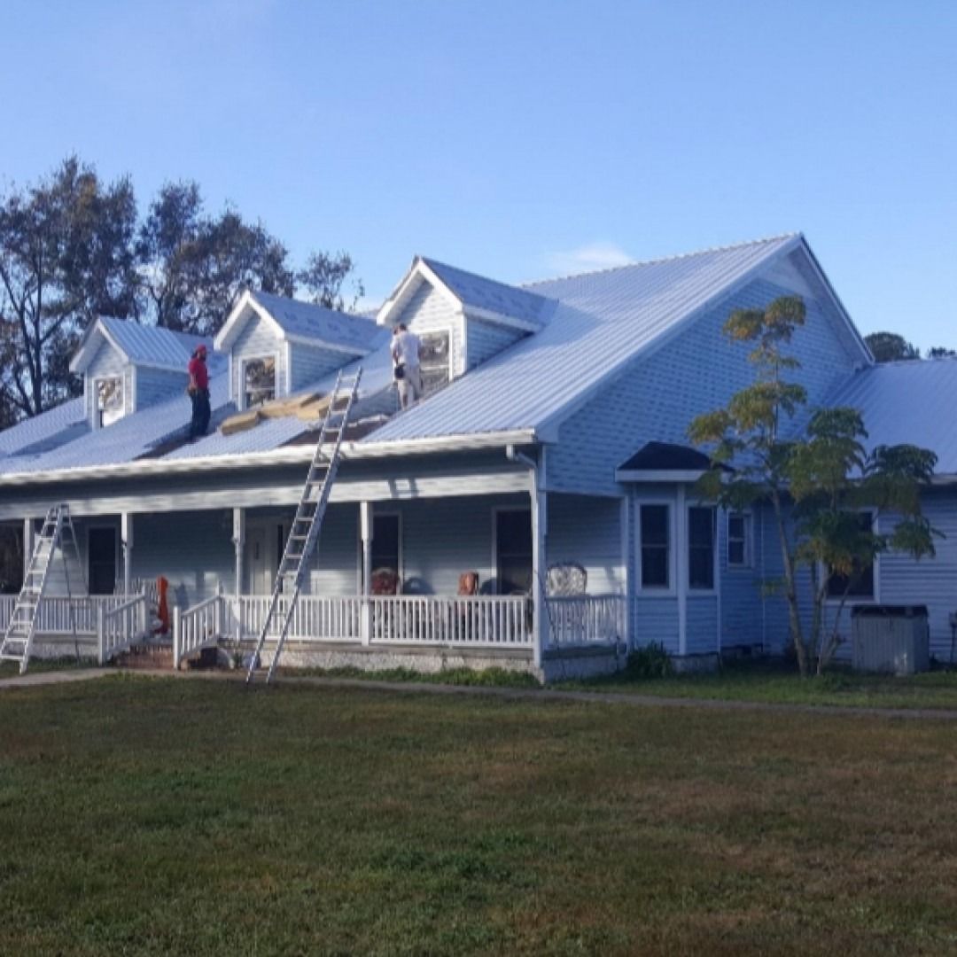 White house with porch and ladders on the roof, appearing under repair in a grassy yard.