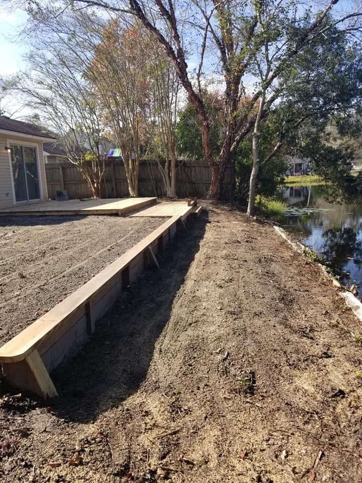 Backyard with a long wooden deck beside a dirt yard and a narrow water canal, trees in the background