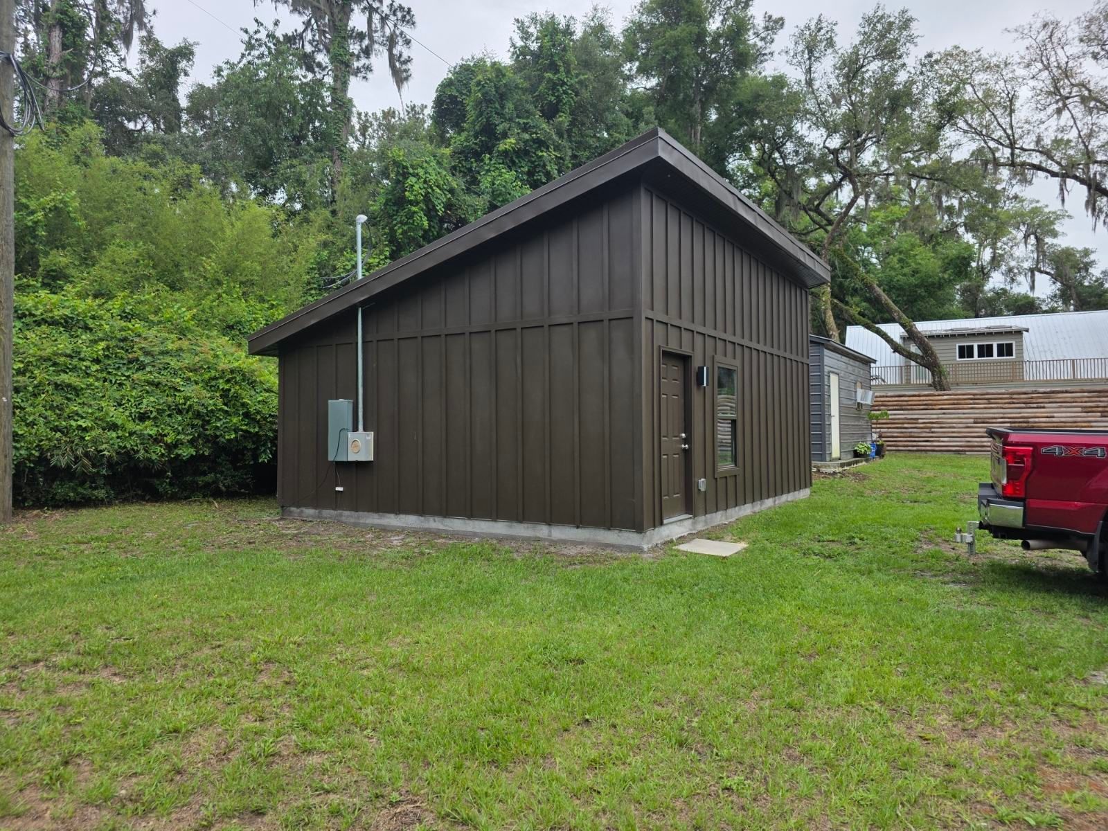 Dark brown shed with a gabled roof on a grassy yard, with trees and a red truck nearby.