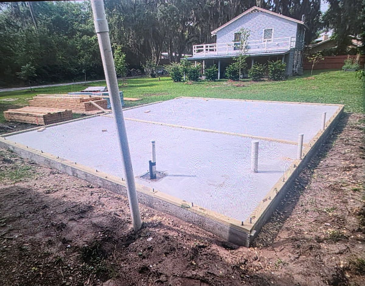 Concrete foundation slab under construction beside a house, with utility pipes and wooden forms visible.