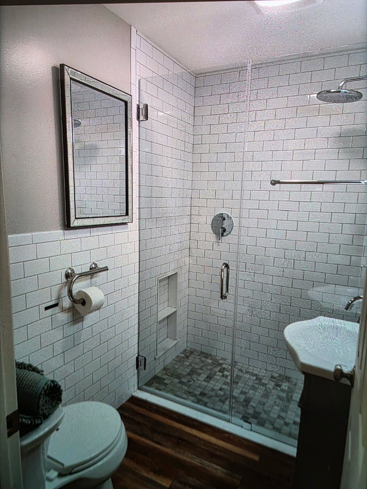 Small white-tiled bathroom with a glass-enclosed shower, toilet, sink, and wall mirror.