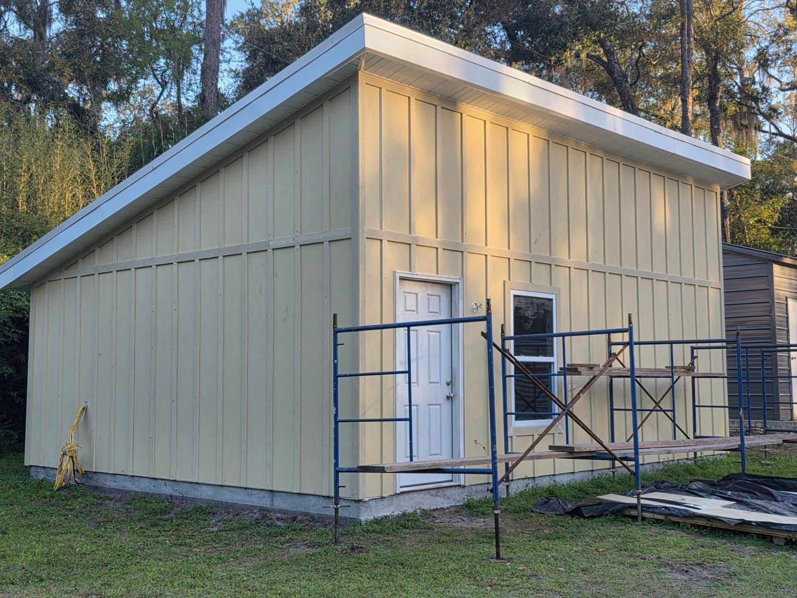 Beige shed under construction with scaffolding and a white door beside trees.