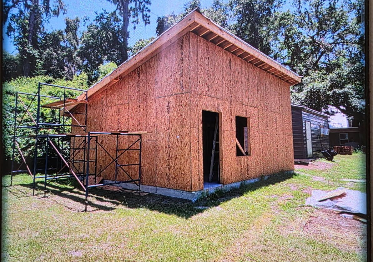 Partially built wooden house with OSB siding and scaffolding in a grassy yard, trees in the background