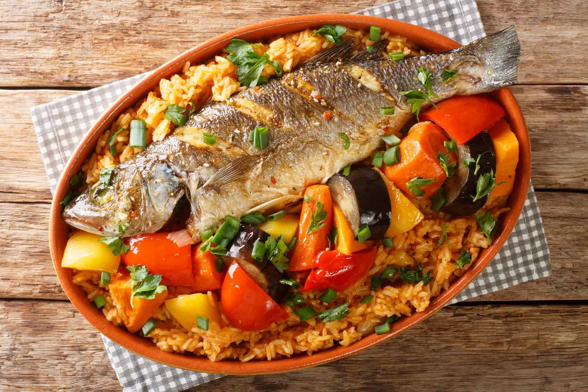 Roasted fish and vegetables in a clay dish on a checkered napkin, set on a wooden table.