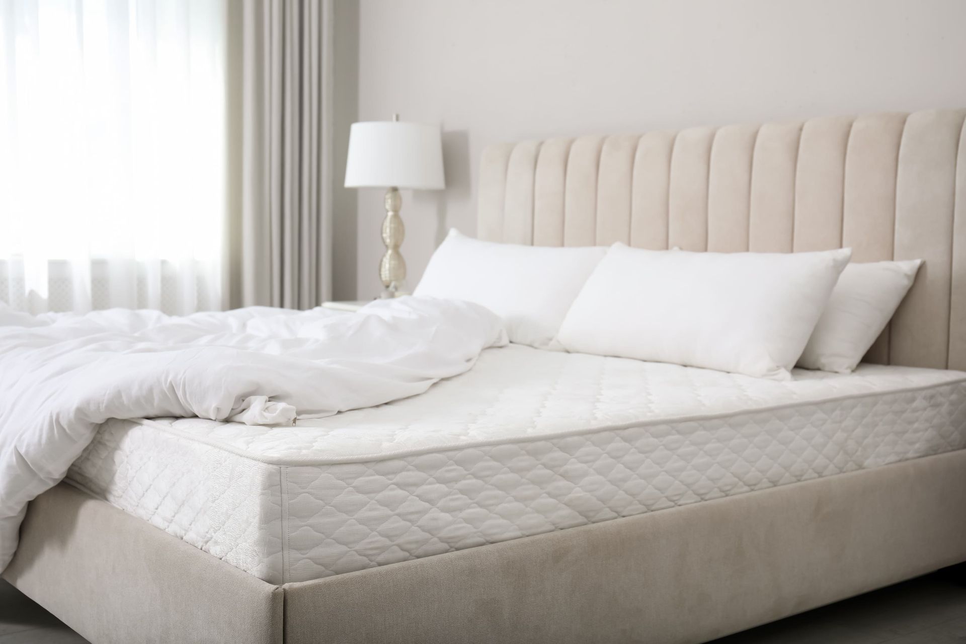 Beige bed with white bedding, pillows, and a lamp in a bright bedroom setting.